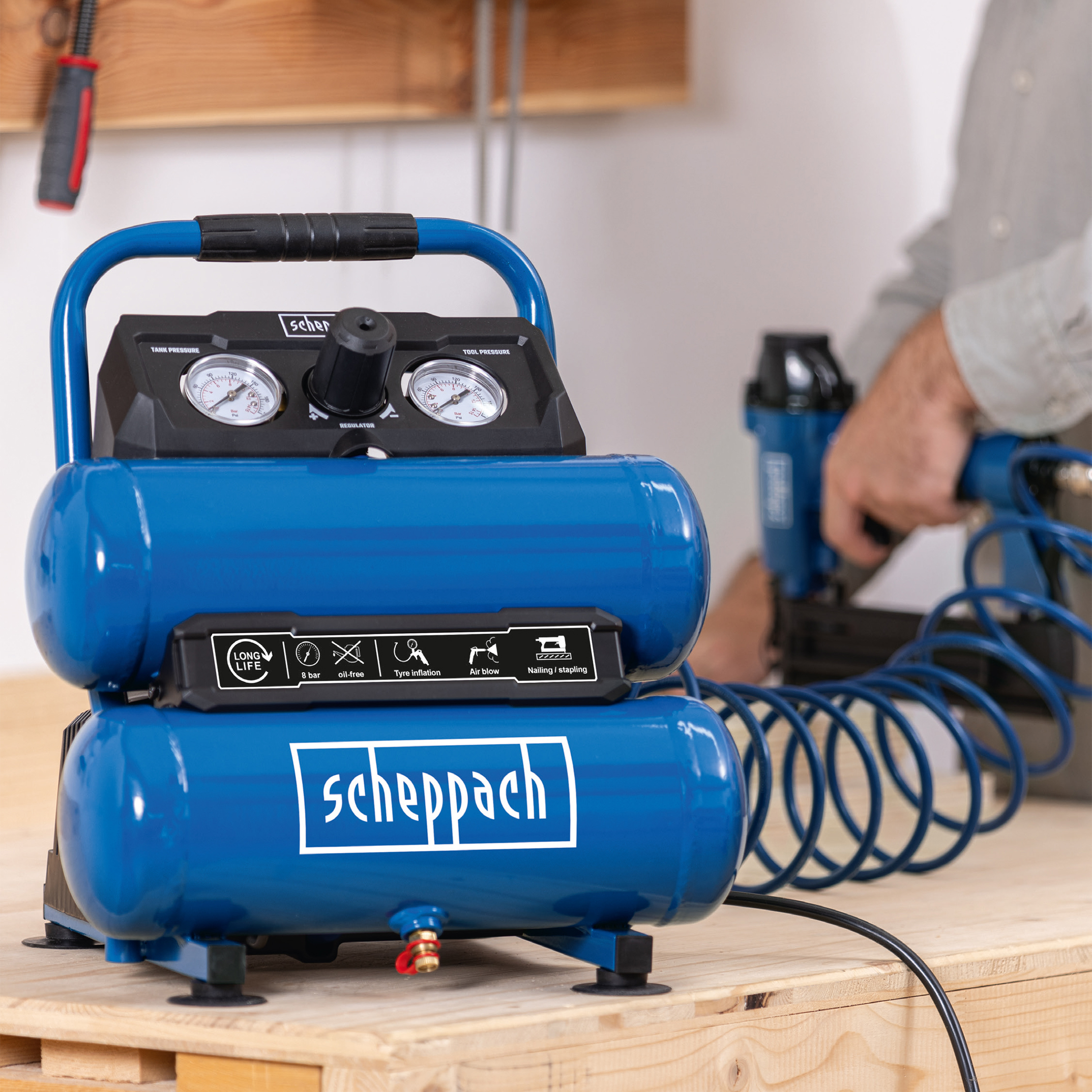 Blauwe luchtcompressor op een houten tafel, met een persoon die een tacker gebruikt op de achtergrond.