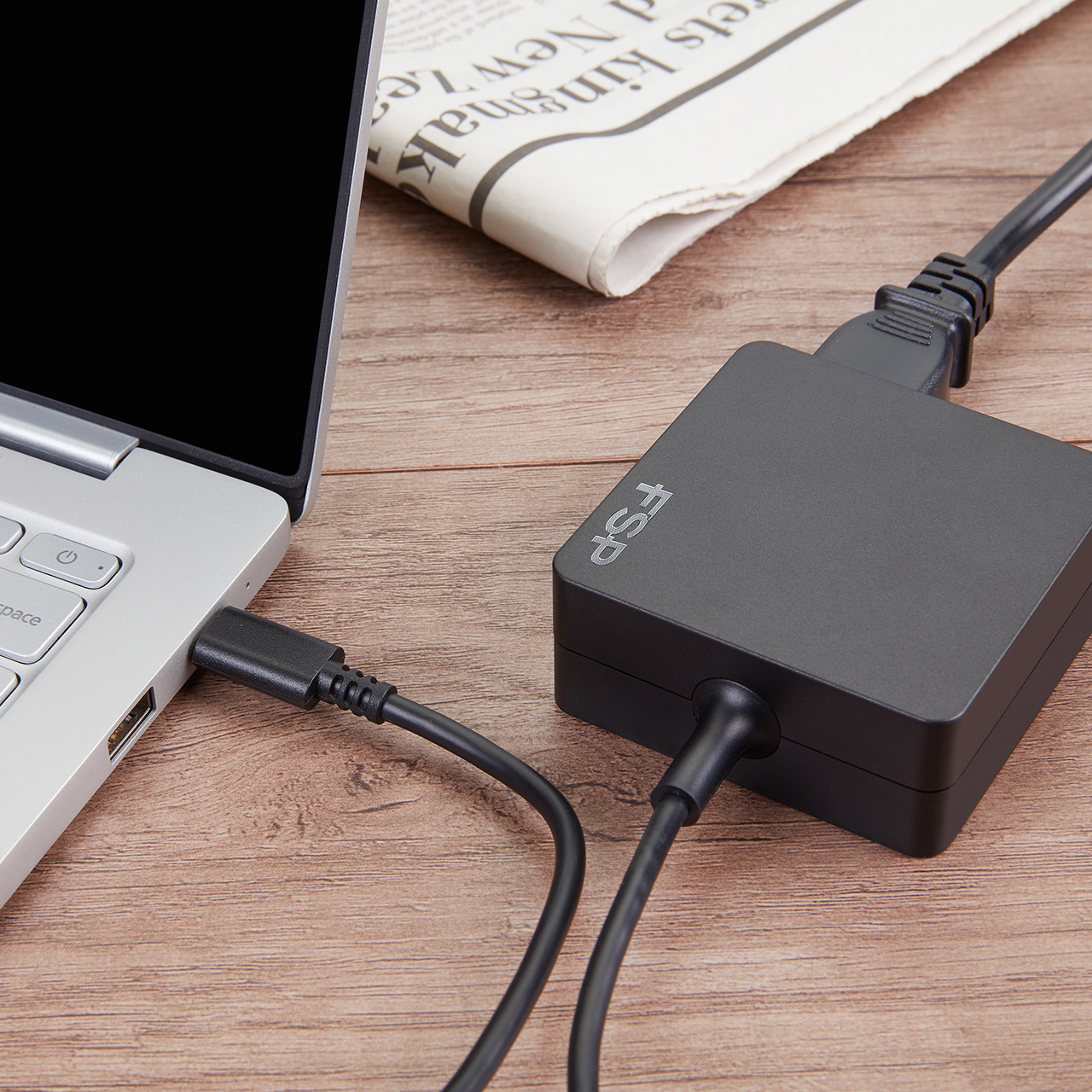 Laptop, der von einem schwarzen FSP-Adapter aufgeladen wird. Adapter und Kabel liegen auf Holz.