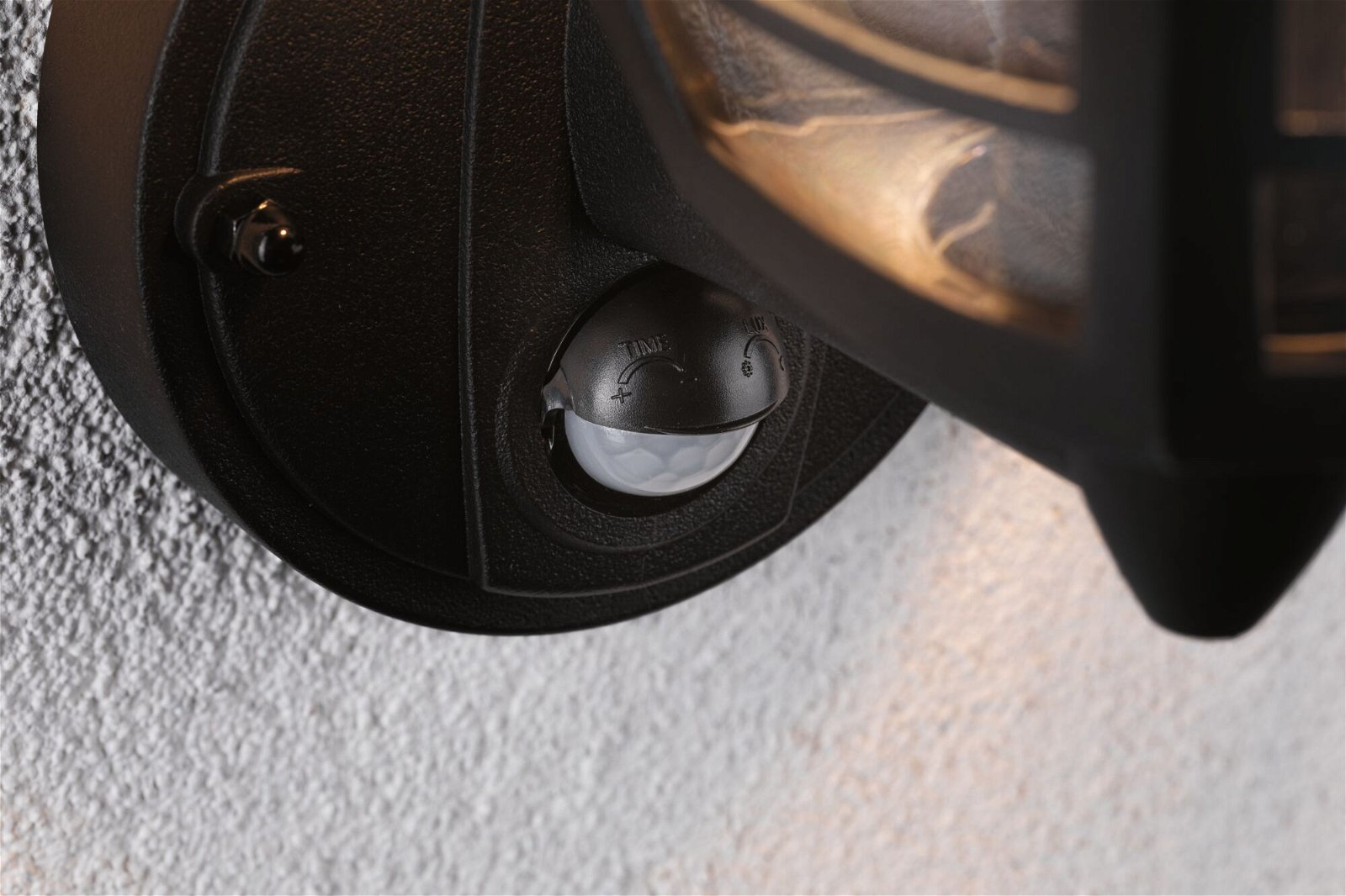 Czarna zewnętrzna lampa ścienna z czujnikiem ruchu, na białej ścianie.