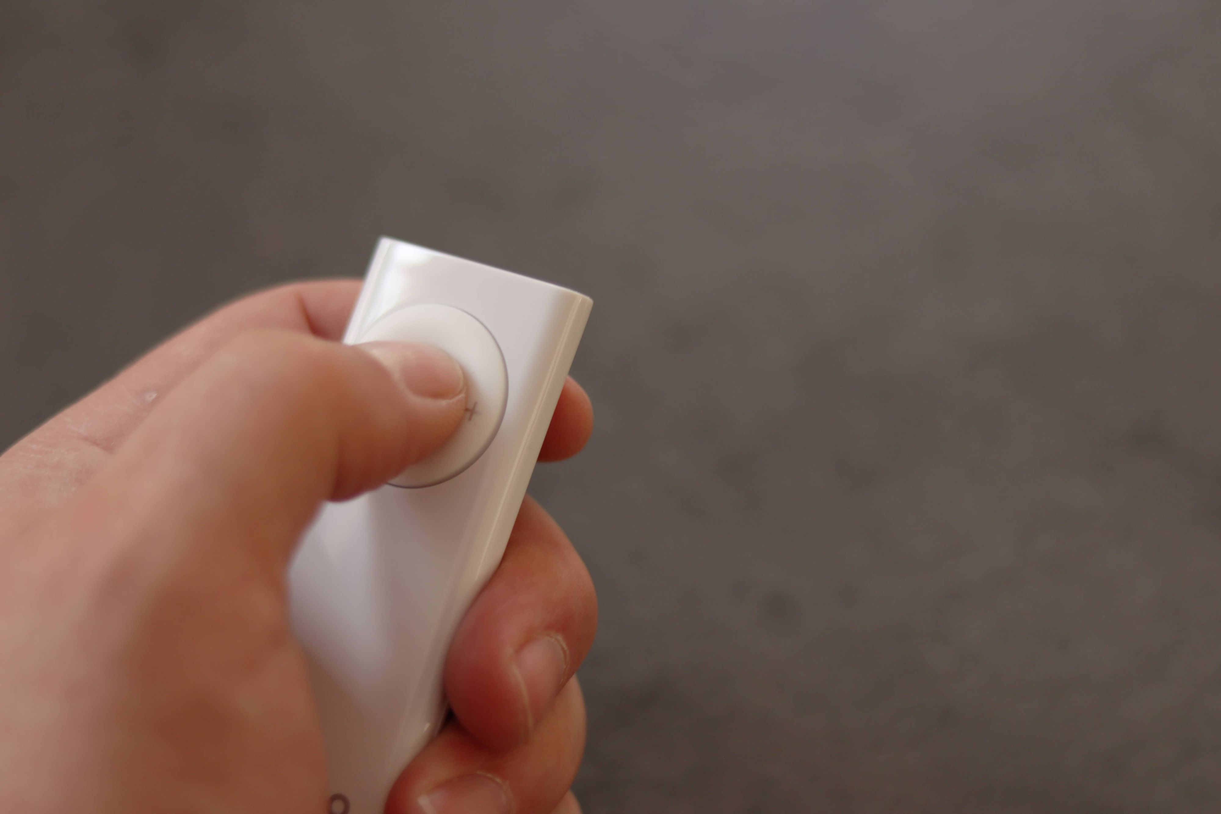 Hand hält eine weiße Fernbedienung mit rundem Knopf. Dunkler Hintergrund.