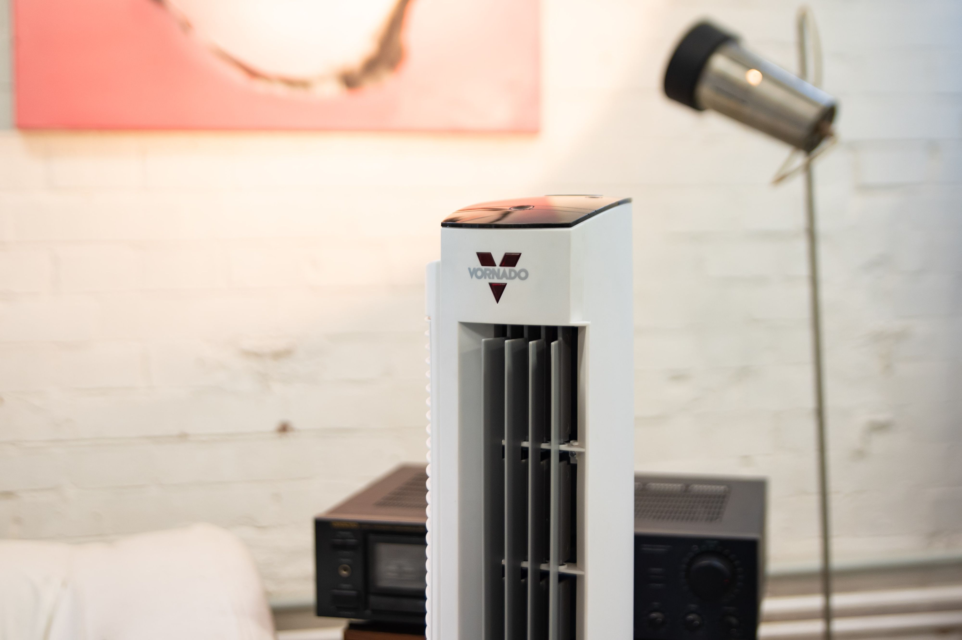 Weißer Vornado-Ventilator mit Logo. Hintergrund: rosa Gemälde, Holzwand, schwarze Stereoanlage.