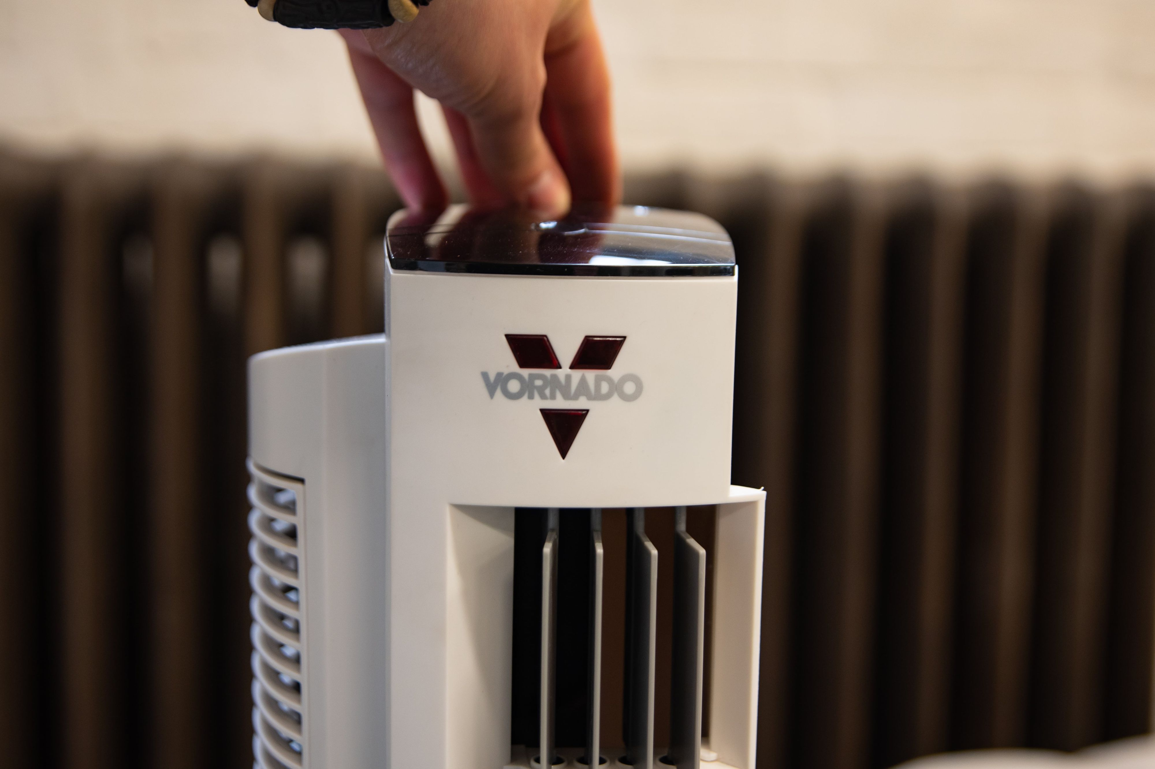 Hand drückt Knopf an weißem Vornado-Ventilator. Vornado-Logo sichtbar. Brauner Heizkörper.