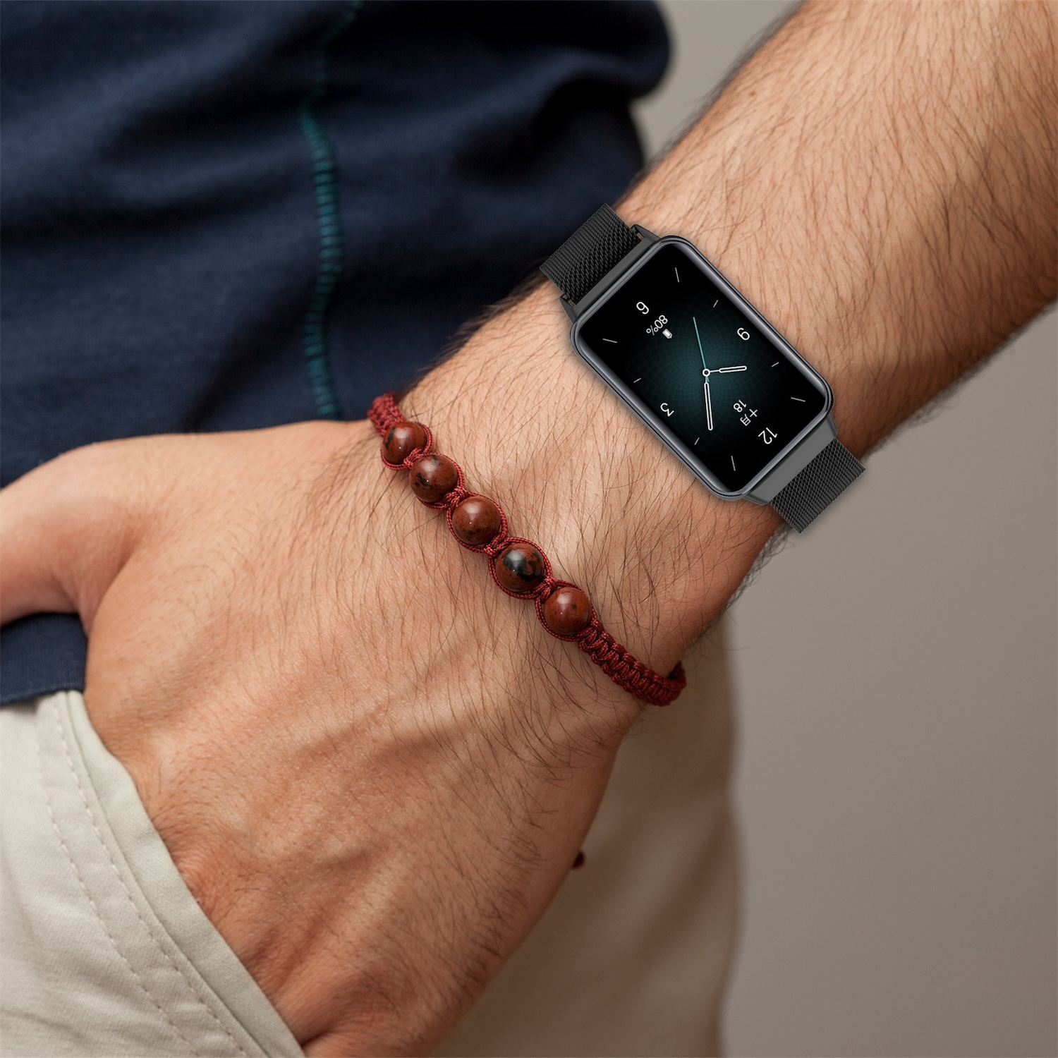 Das Handgelenk eines Mannes mit einer schwarzen Smartwatch und einem roten Perlenarmband. Die Uhr zeigt die Uhrzeit an.