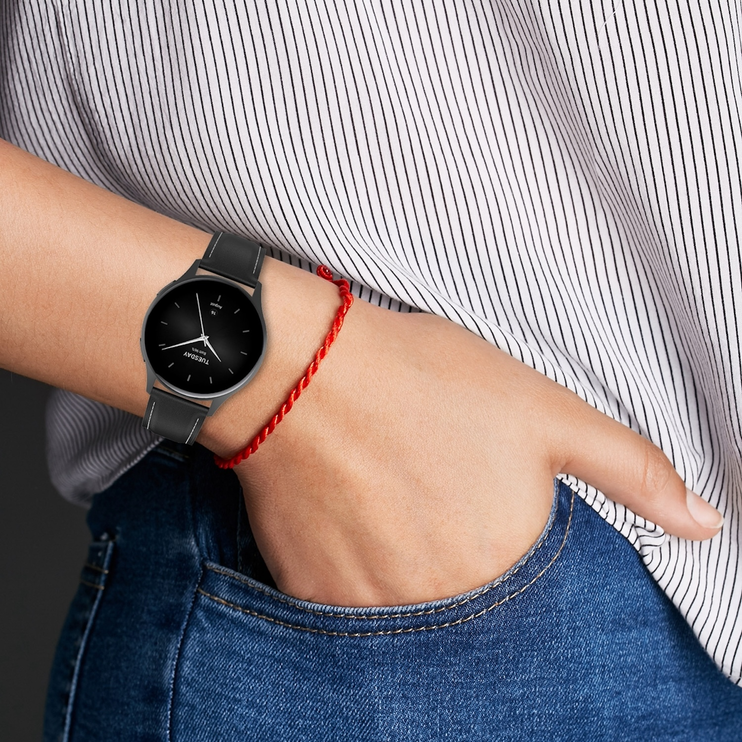 Das Handgelenk einer Person mit einer schwarzen Smartwatch und einem roten Faden. Die Person trägt ein weißes Hemd und Jeans.