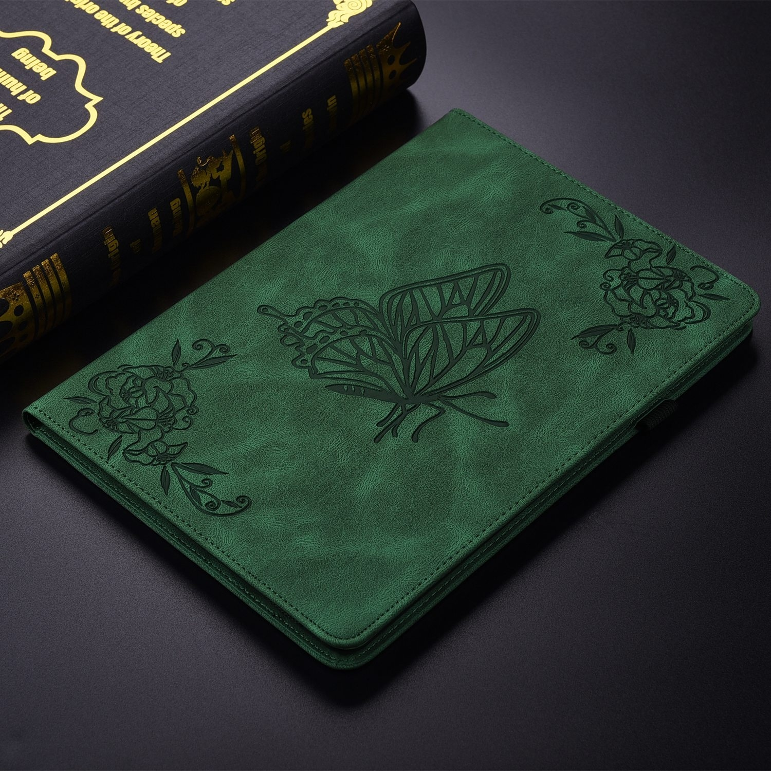 Funda verde para tableta con diseño de mariposa y adornos florales. Un libro negro cerca.