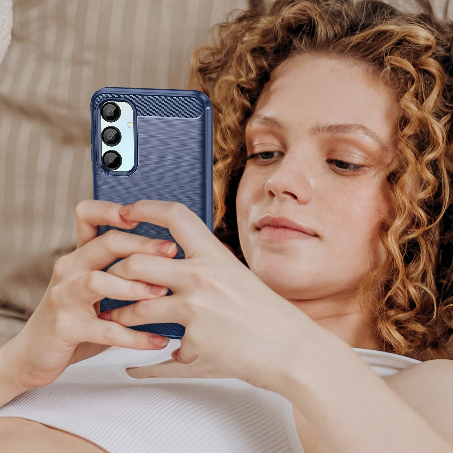 Frau hält blaues Telefon mit blauer Hülle, liegt im Bett. Lockiges Haar und weißes Oberteil sichtbar.
