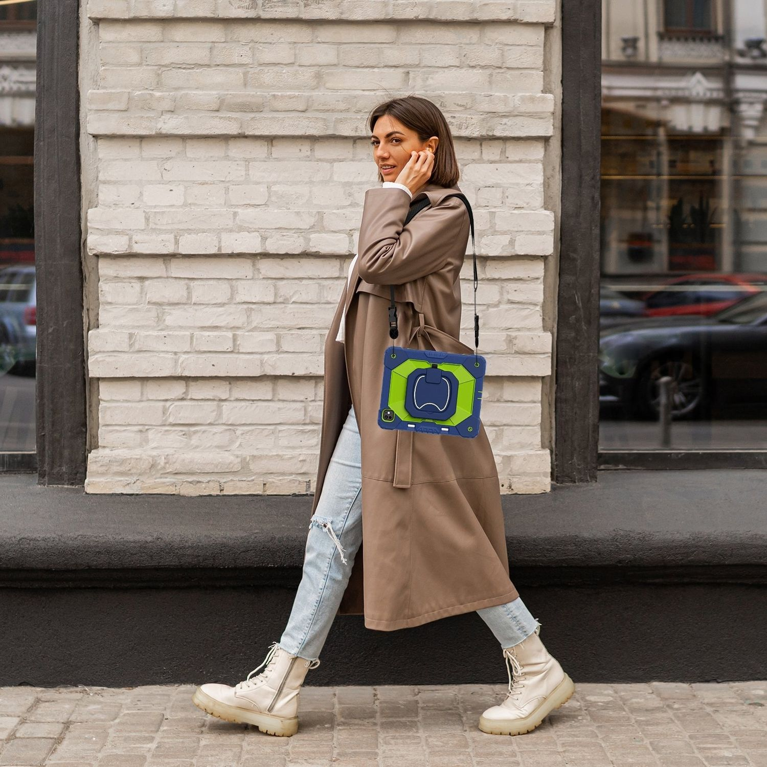 Mujer camina, con abrigo marrón, llevando una funda para tablet con correa para el hombro, sobre ladrillo y calle.