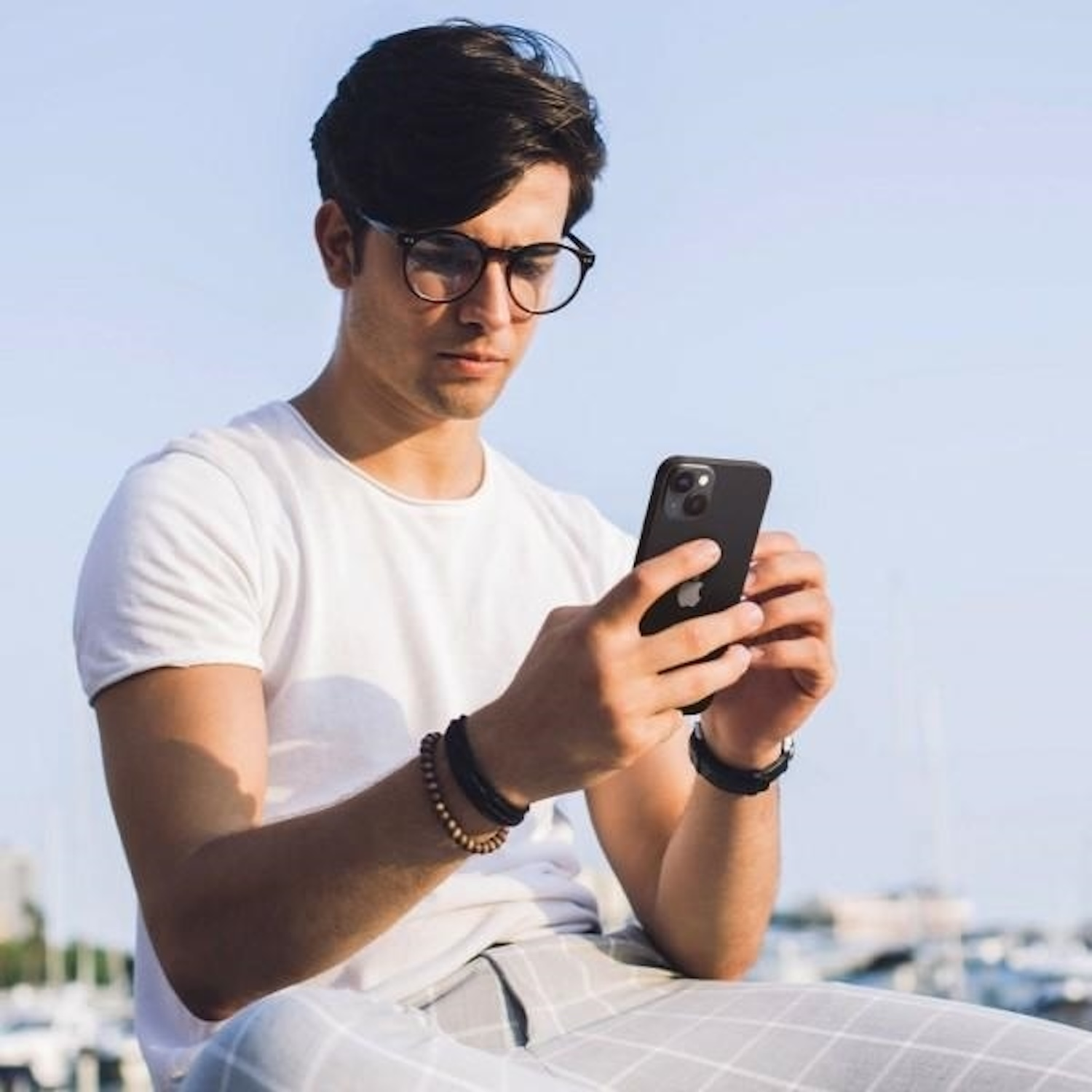 Man in white shirt and glasses looks at his phone. Blue sky background.