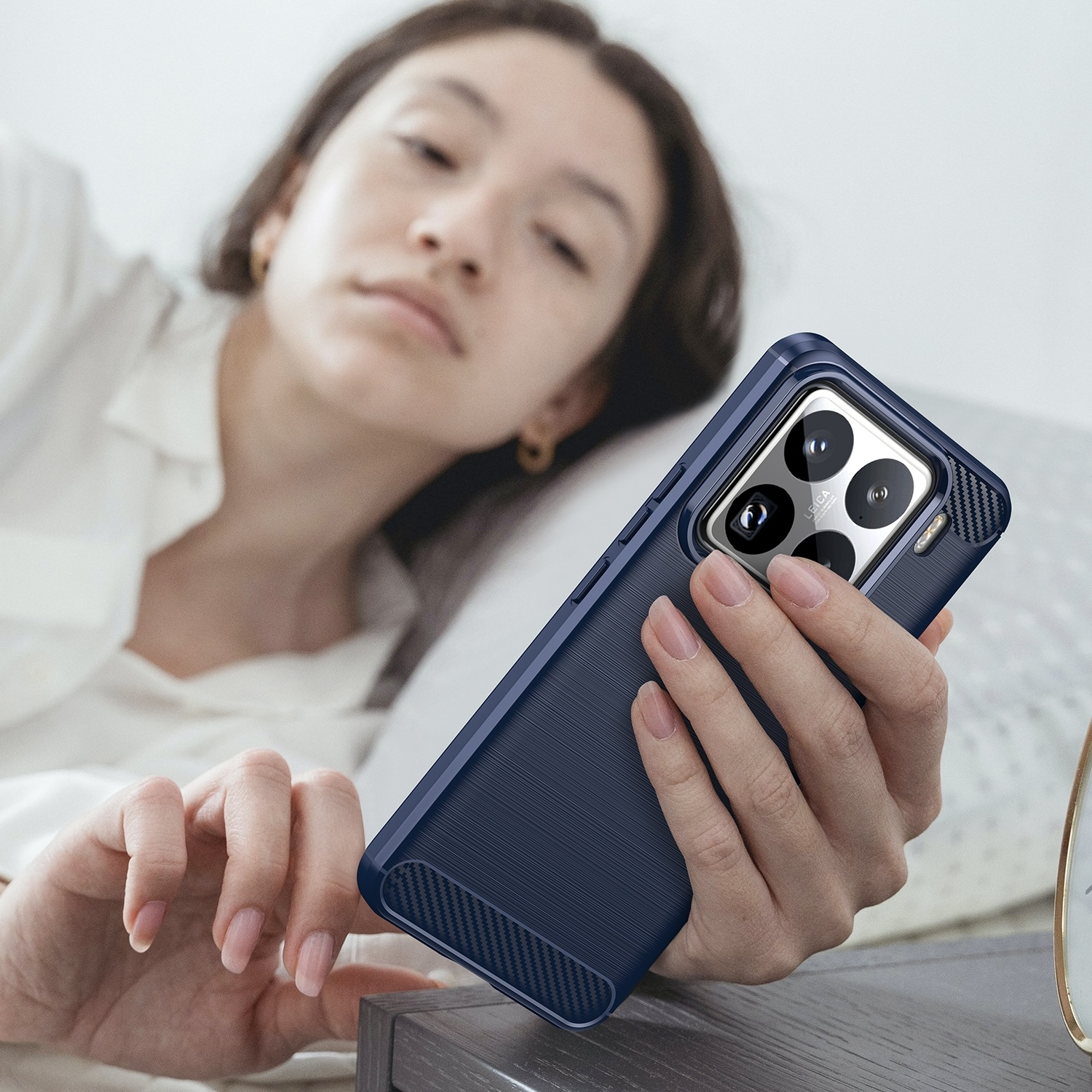 Frau hält ein dunkelblaues Telefon mit Hülle. Sie liegt im Bett und schaut auf das Telefon. Ohrringe sichtbar.