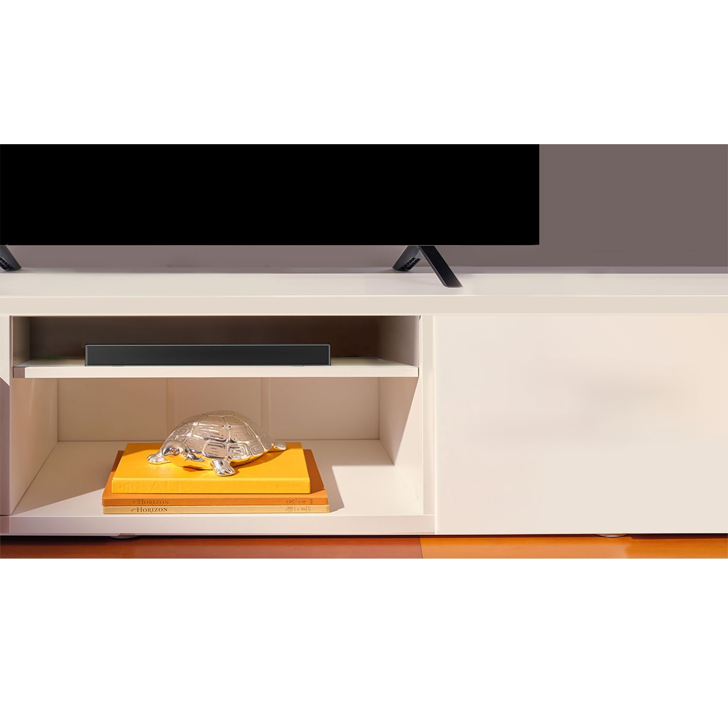 Een tv staat op een witte tv-kast. Een soundbar staat op de plank. Decoratieve items, waaronder boeken, zijn zichtbaar.