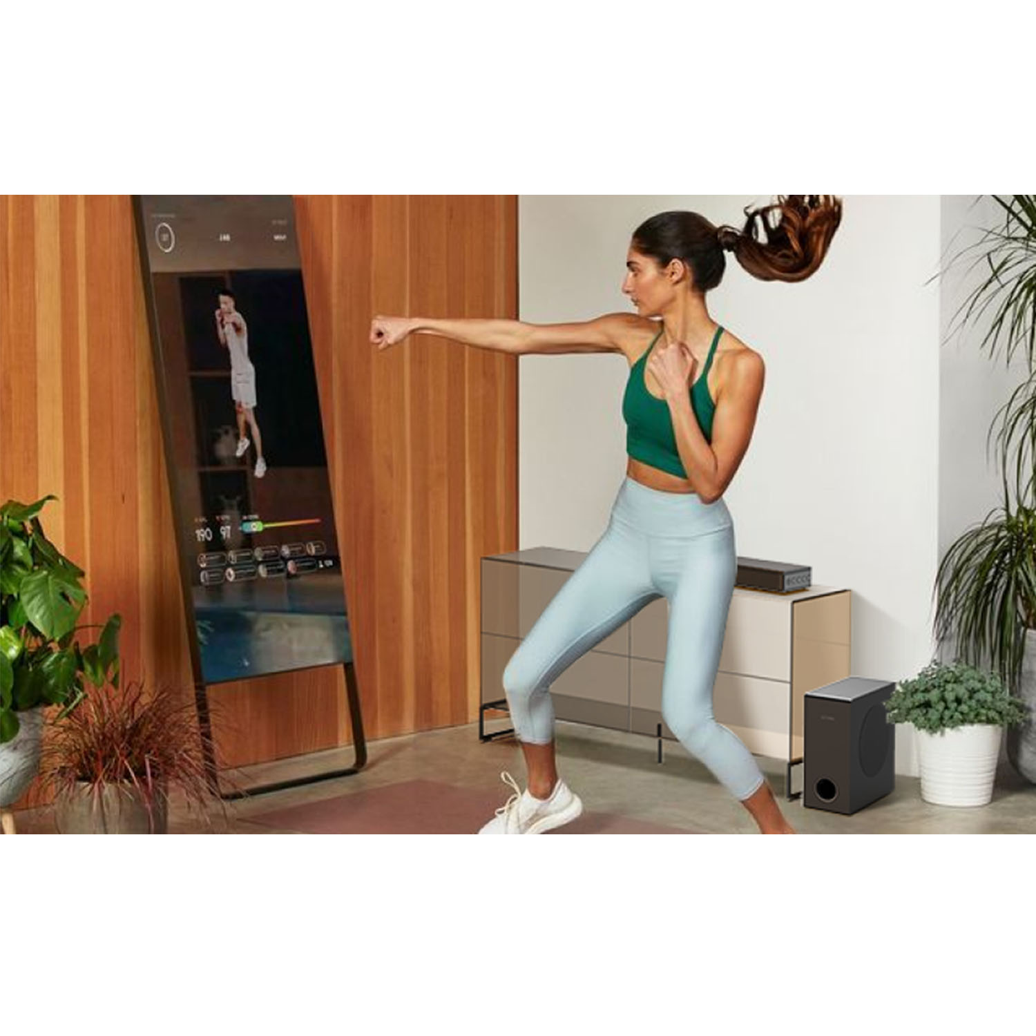 Vrouw bokst voor een slimme spiegel. Een subwoofer staat rechts. Houten muur en een plant in de hoek.