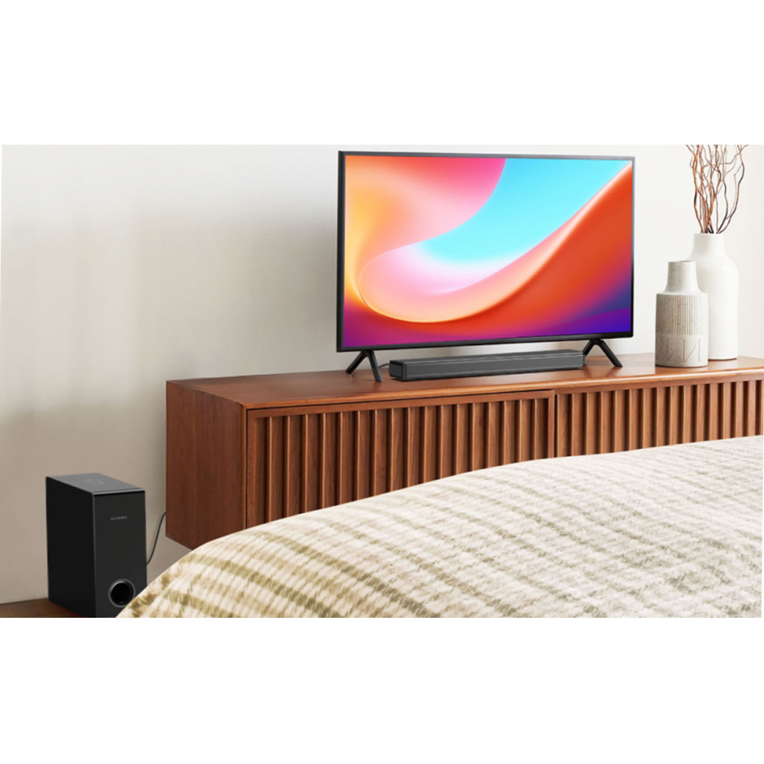 TV op houten kast, soundbar, subwoofer, vaas met takjes en een bed met een patroon deken.