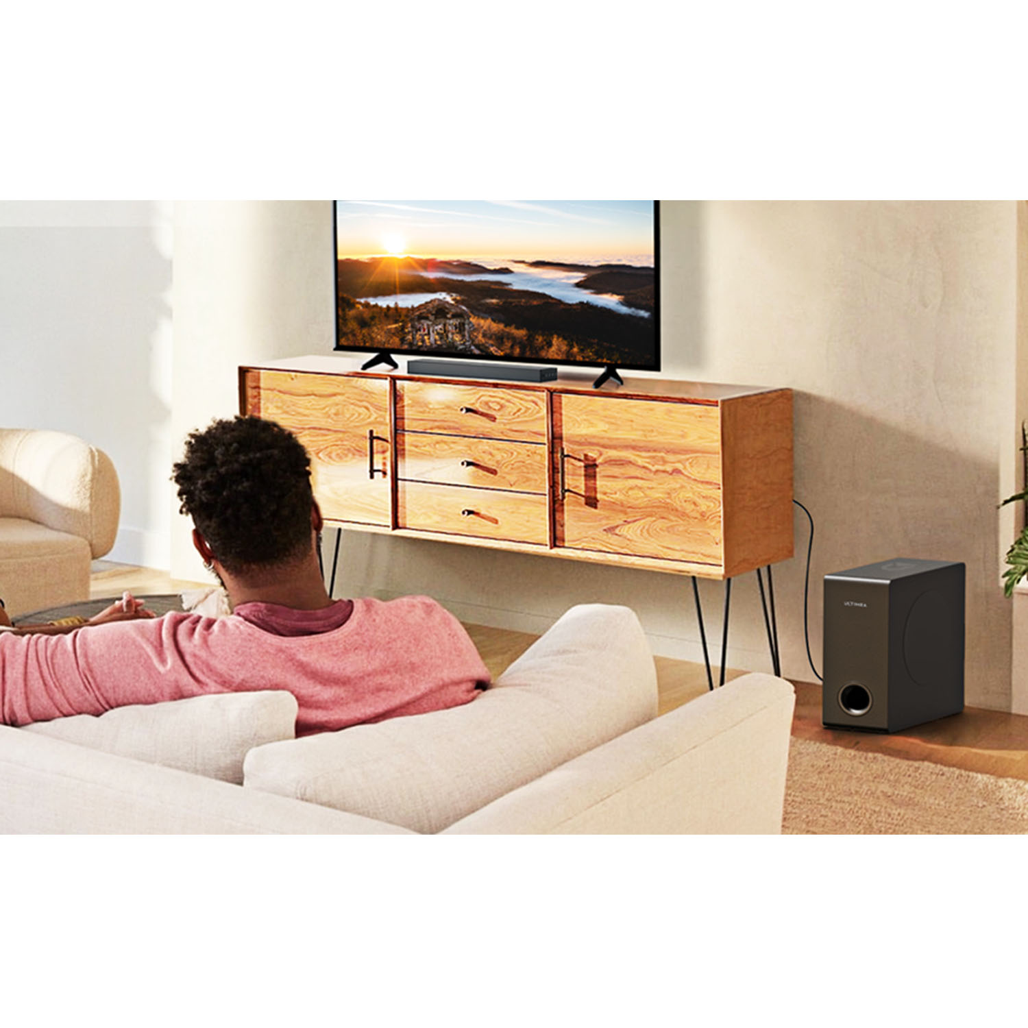 Een persoon kijkt tv. Een soundbar staat onder de tv. Een subwoofer staat rechts. Een houten kast.