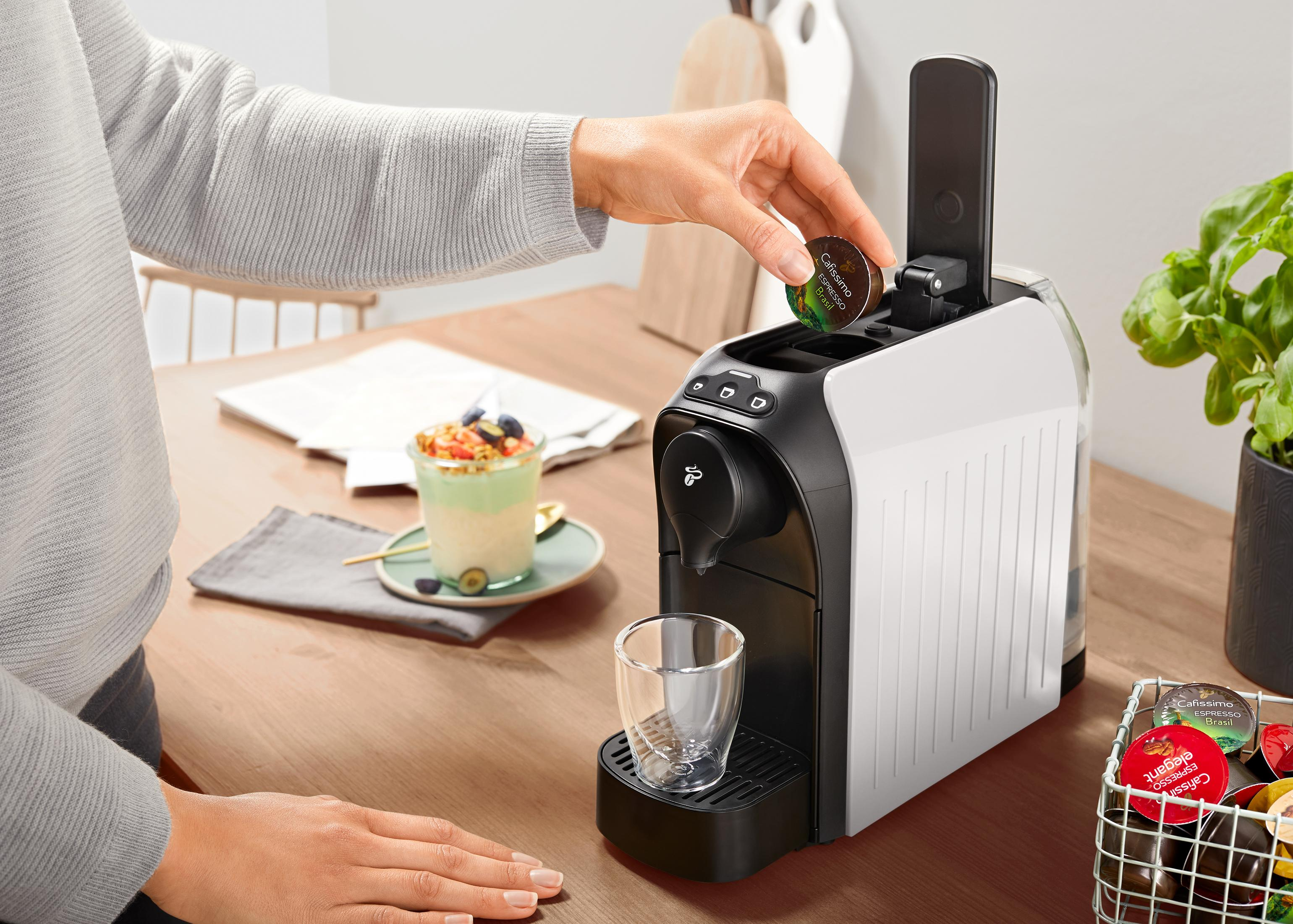 Person fügt Kaffeepad in die Kaffeemaschine ein. Es gibt eine Tasse und Essen.
