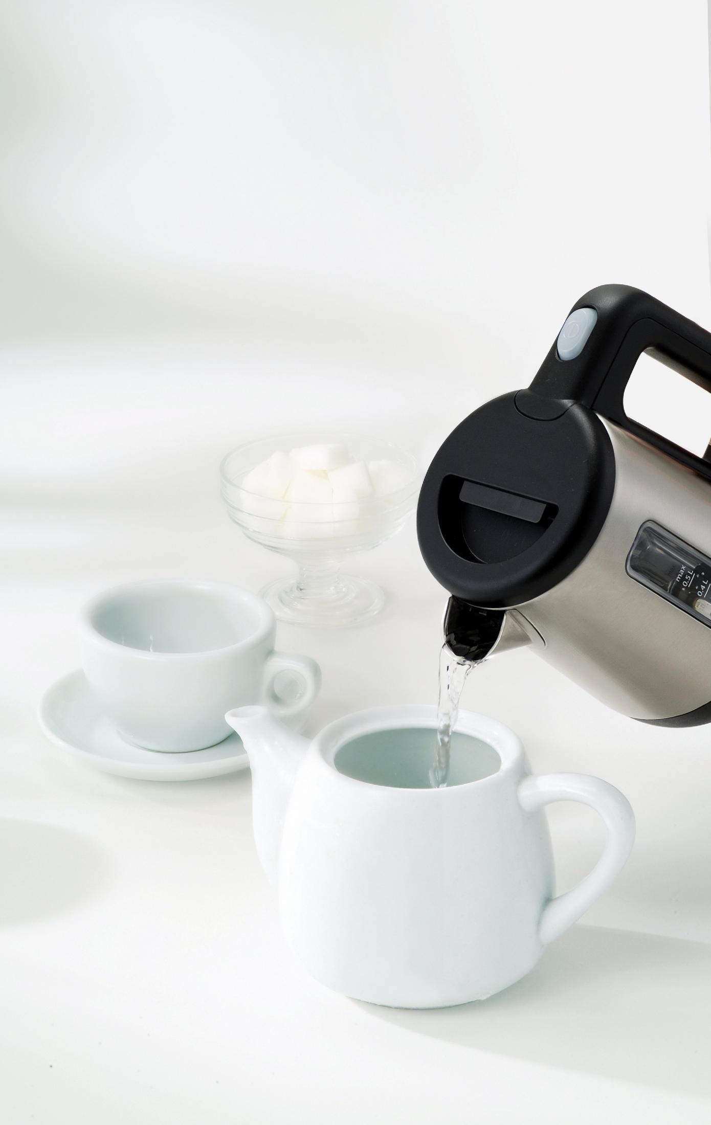 Osoba nalewa wodę ze srebrnego czajnika elektrycznego do białego dzbanka do herbaty, z cukrem i filiżanką w pobliżu.