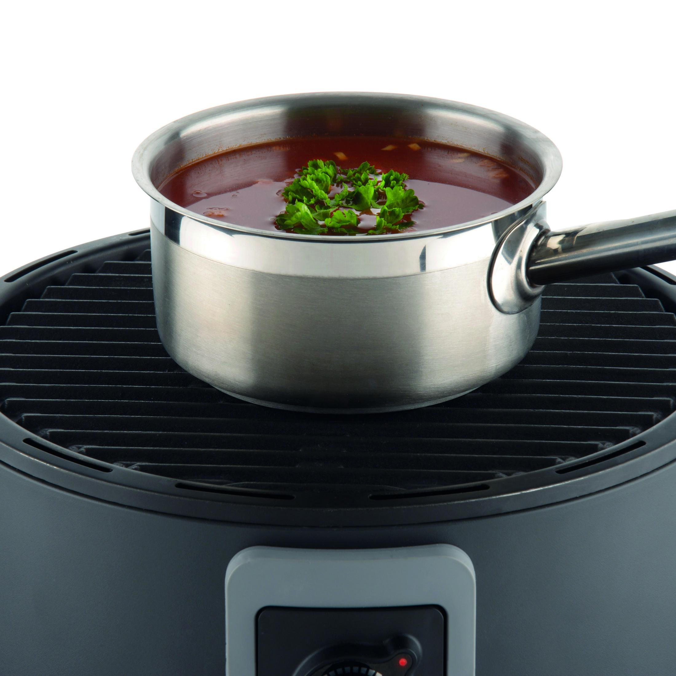 Ein Edelstahltopf mit Suppe und Kräutern steht auf einem schwarzen Grill. Der Grill ist Teil eines runden Geräts.