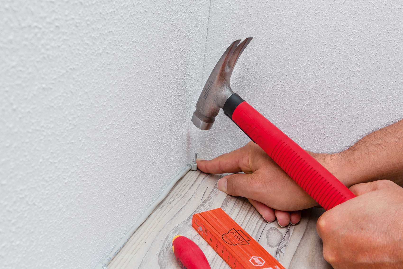 Mano martillando clavo en suelo de madera. Martillo rojo y herramientas visibles.
