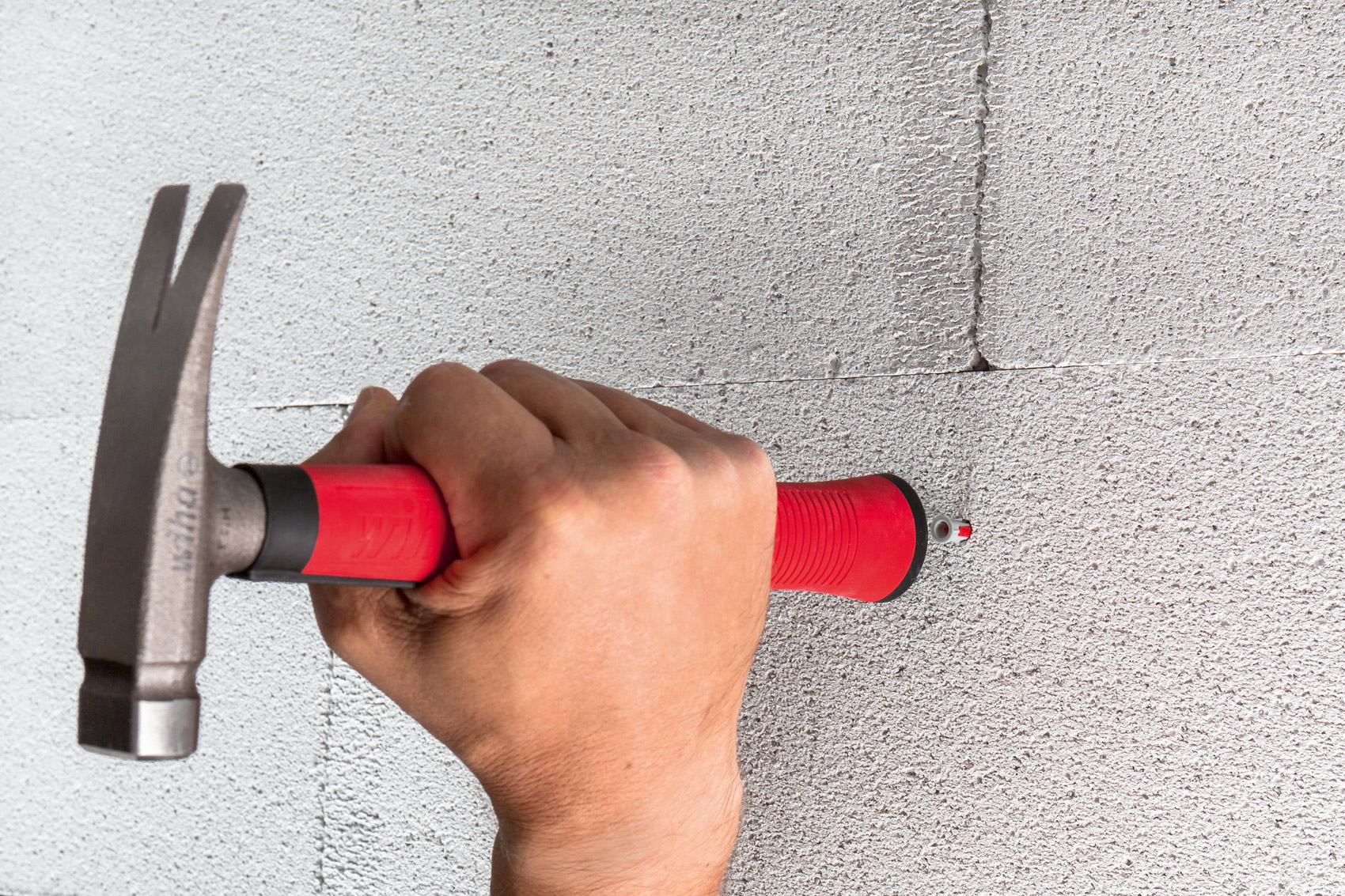 Una mano usando un martillo rojo y plateado para clavar un clavo en una pared de hormigón.