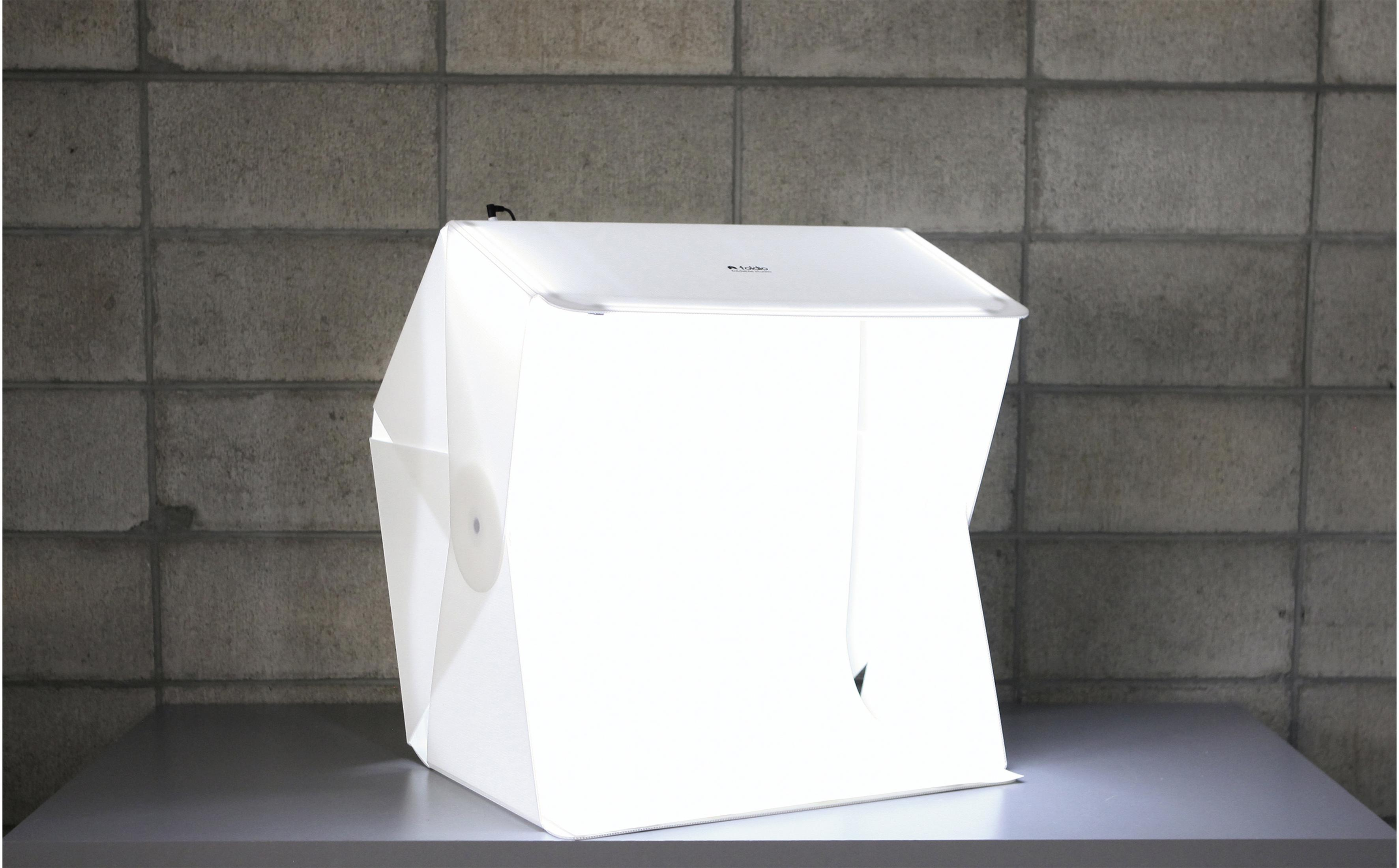 Eine weiße Fotostudiobox auf einer grauen Oberfläche vor einer grauen Ziegelmauer.