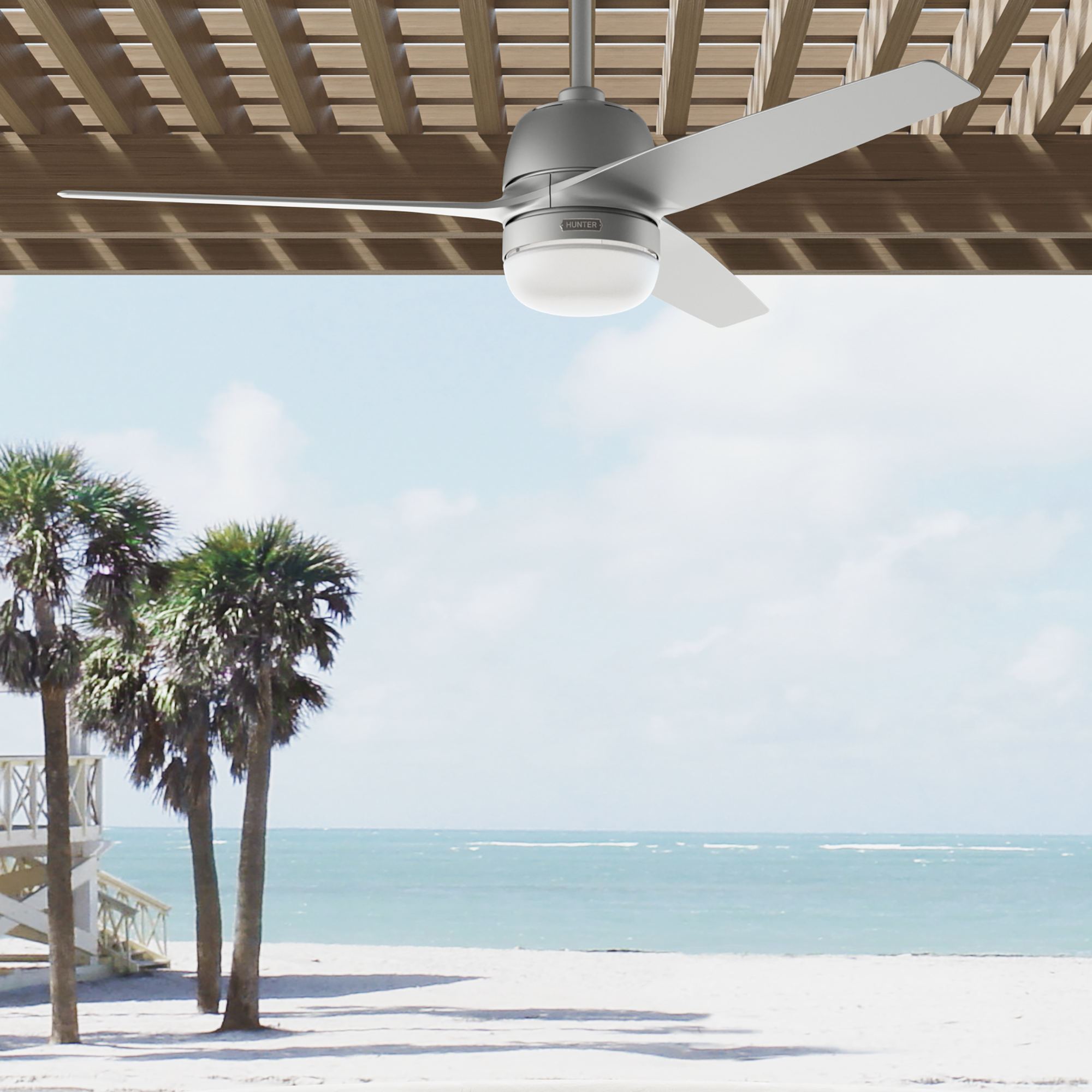 Ein Deckenventilator über einer Strandszene. Palmen, Sand und das Meer sichtbar. Sonniger, blauer Himmel.