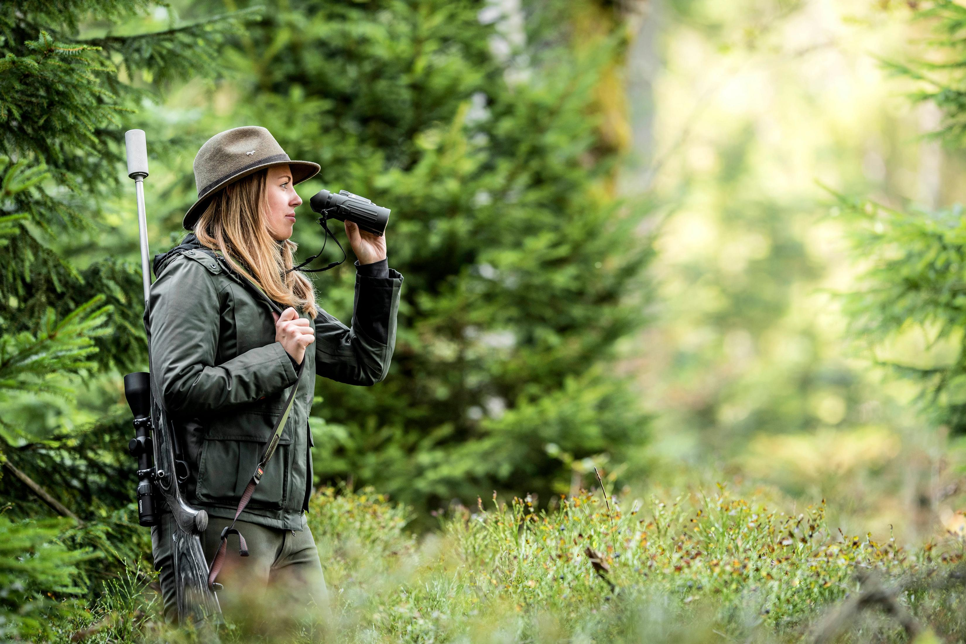 Mujer en bosque con prismáticos y rifle. Árboles en el fondo.