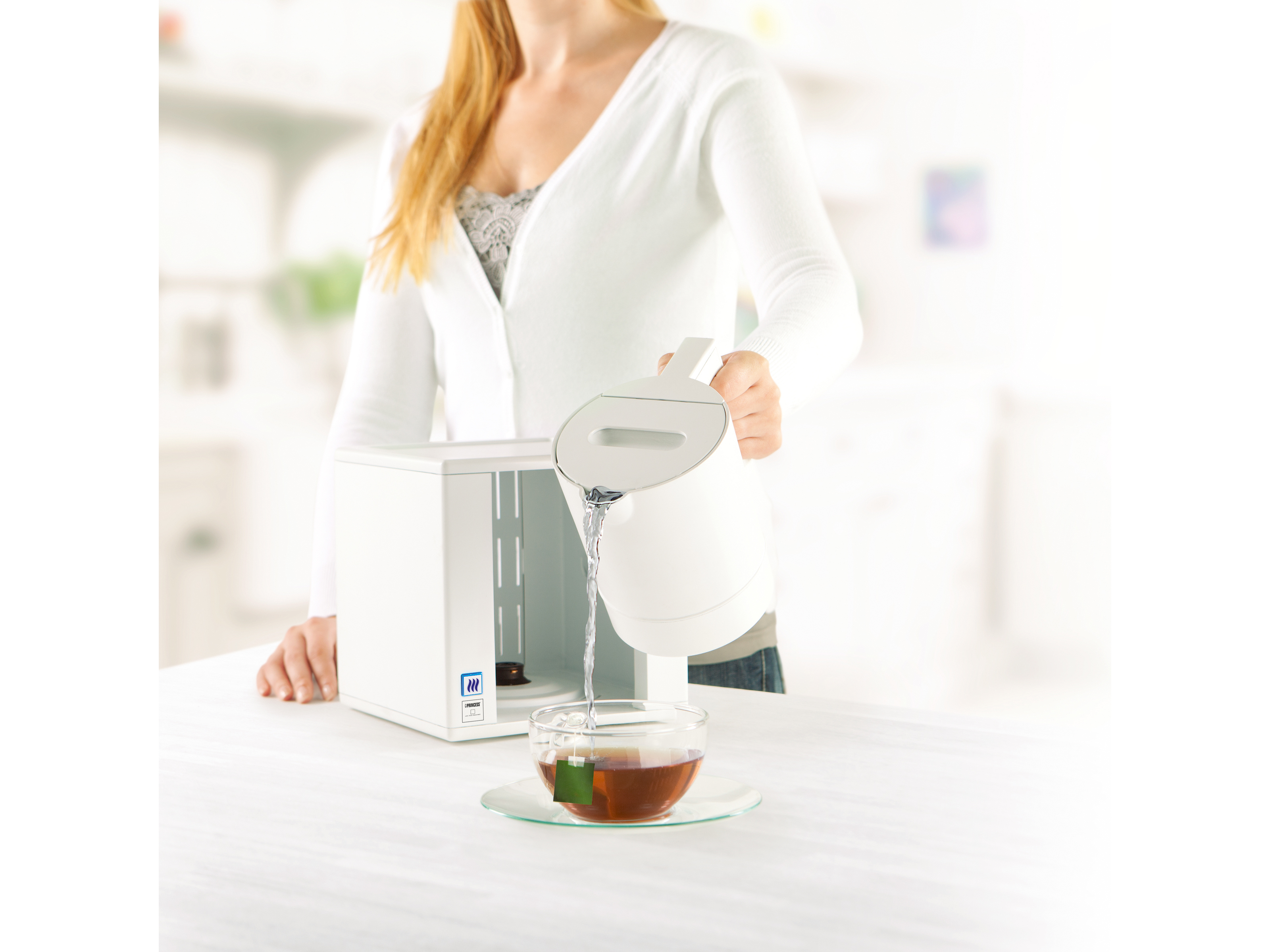 Eine Person gießt Wasser aus einem weißen Wasserkocher in ein klares Glas mit einem Teebeutel auf weißer Oberfläche.
