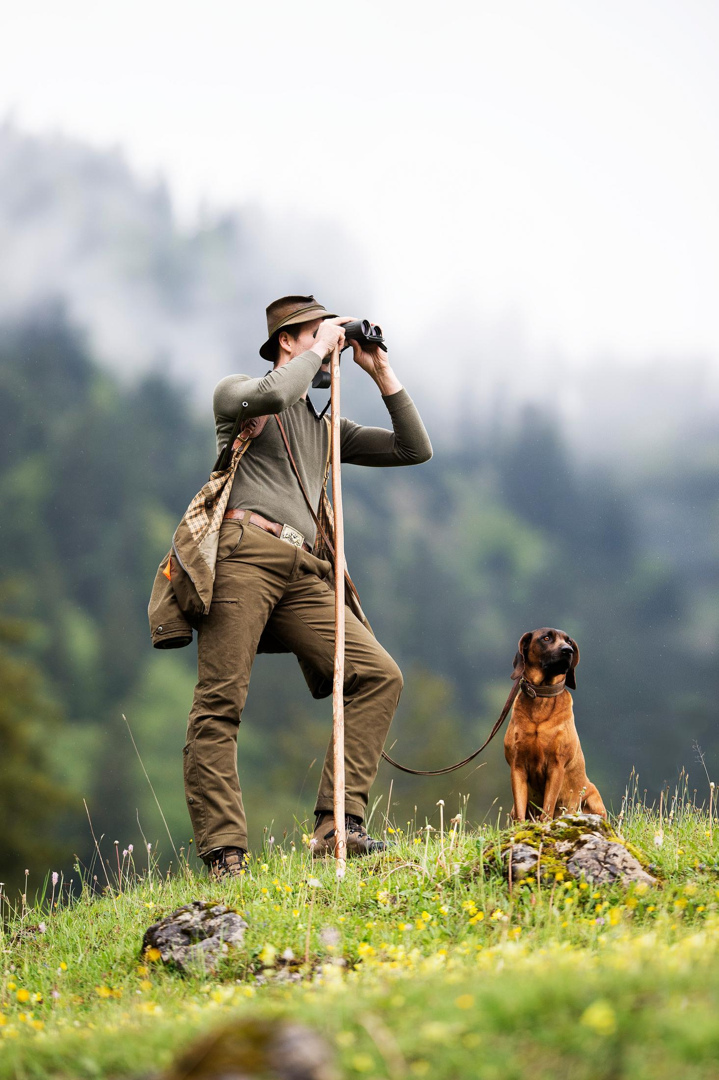 Un hombre con prismáticos y un bastón, observando una escena con un perro.