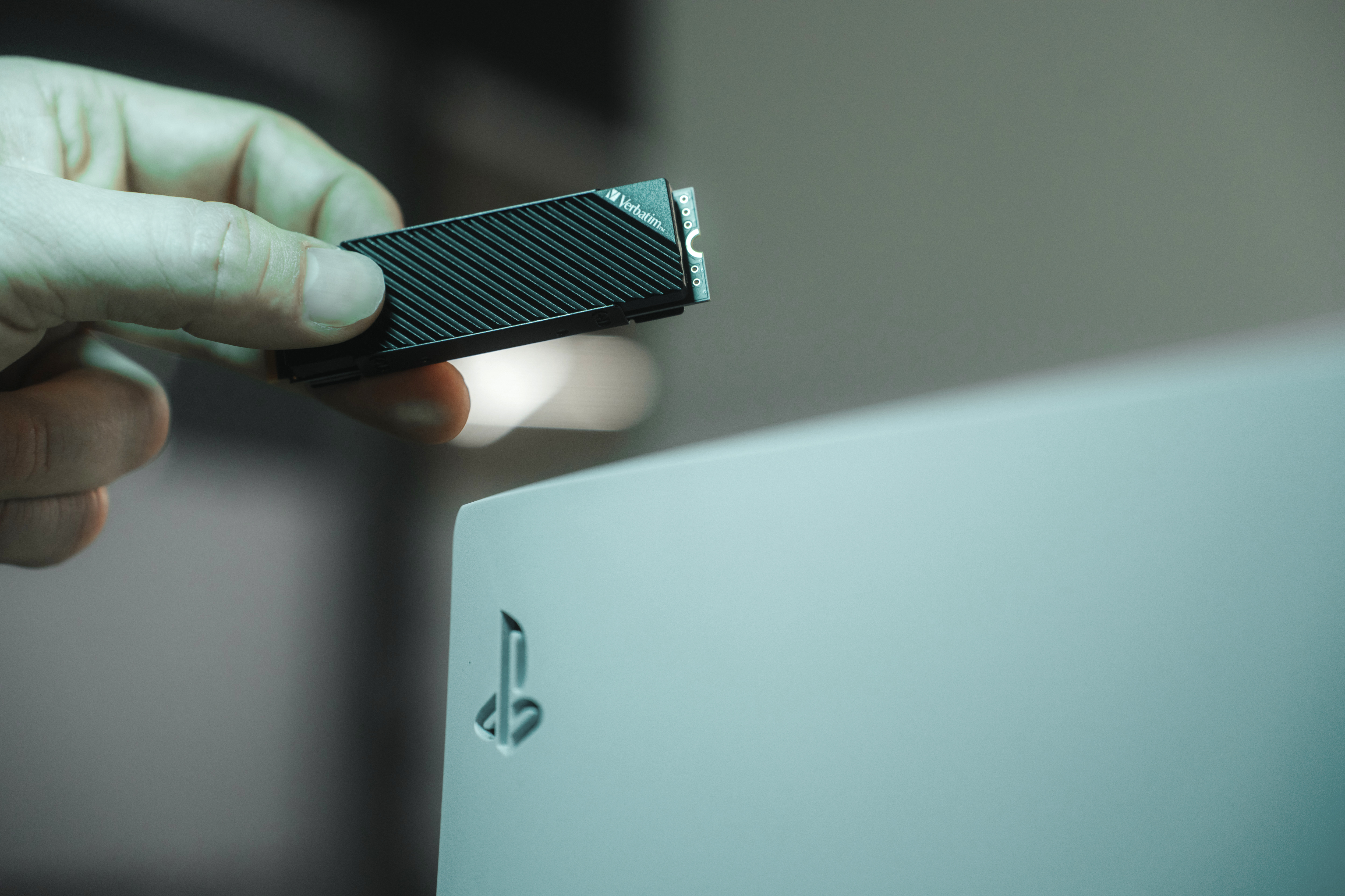 Ręka trzymająca dysk SSD Verbatim, w pobliżu konsoli PlayStation 5.