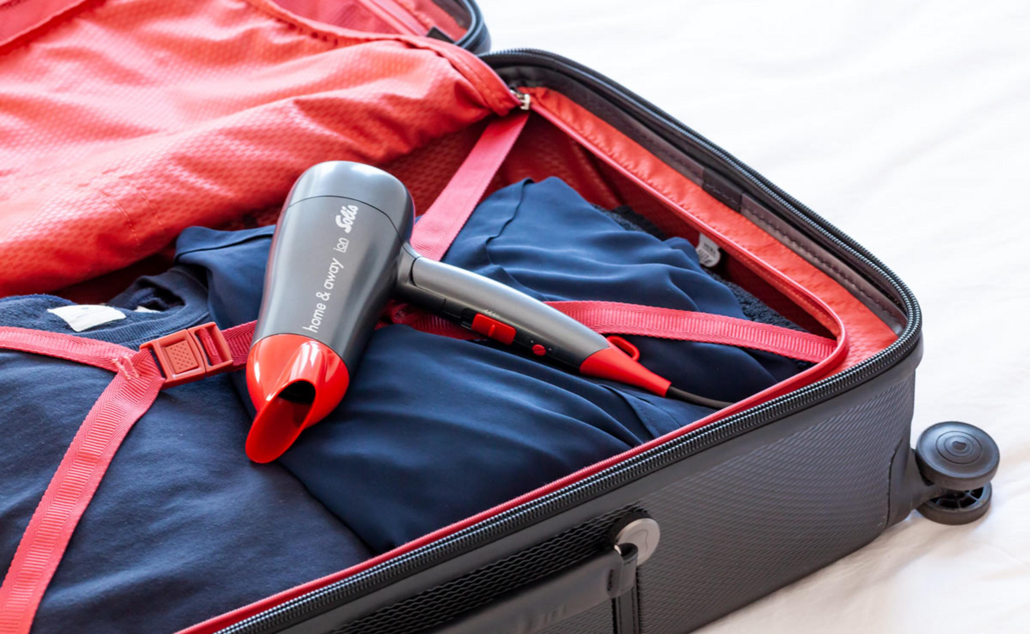 Szara i czerwona suszarka do włosów Solis w otwartej walizce z niebieskimi ubraniami i czerwoną podszewką.