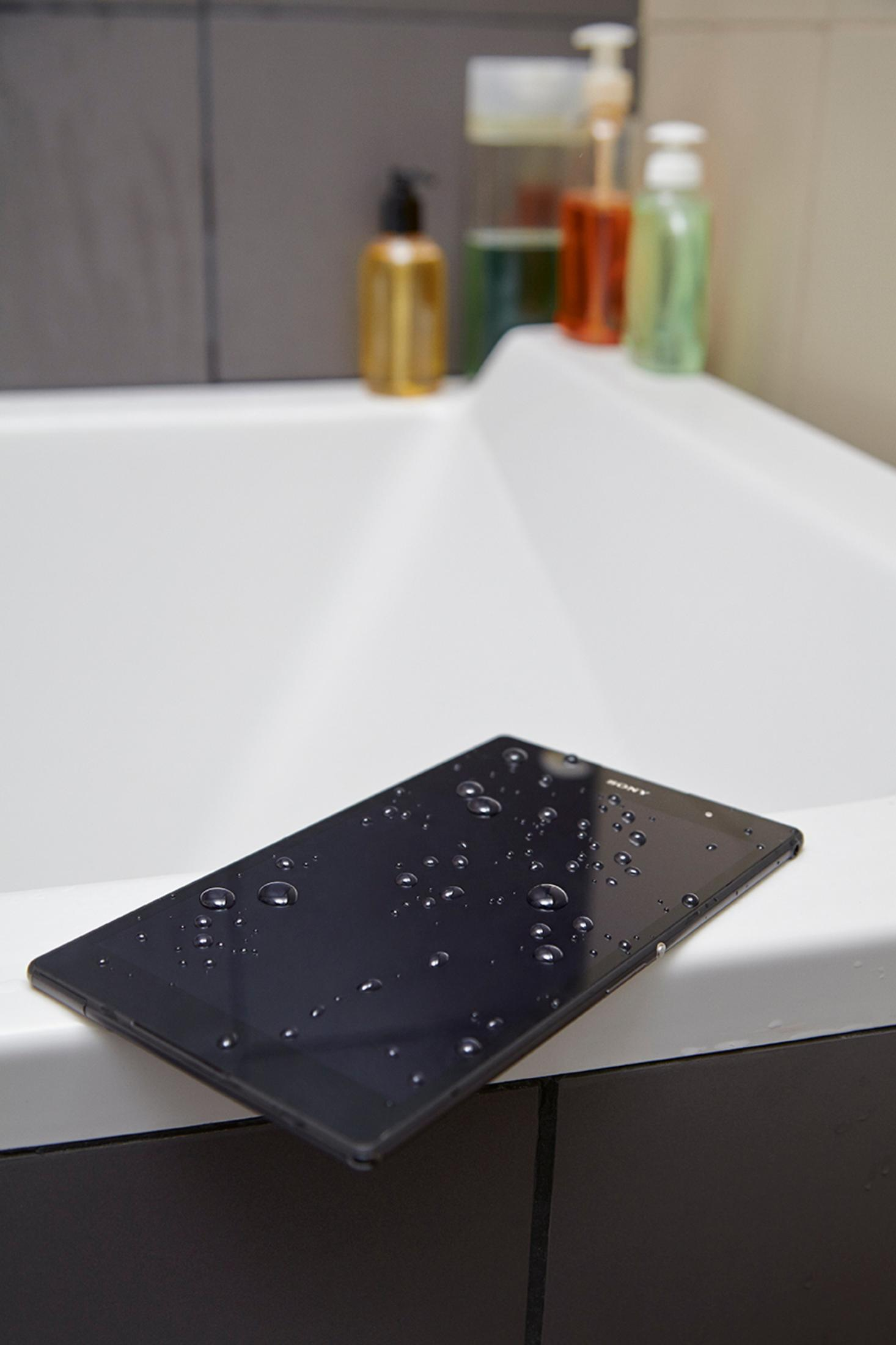 Ein schwarzes Tablet mit Wassertropfen, das auf dem Rand einer weißen Badewanne liegt.