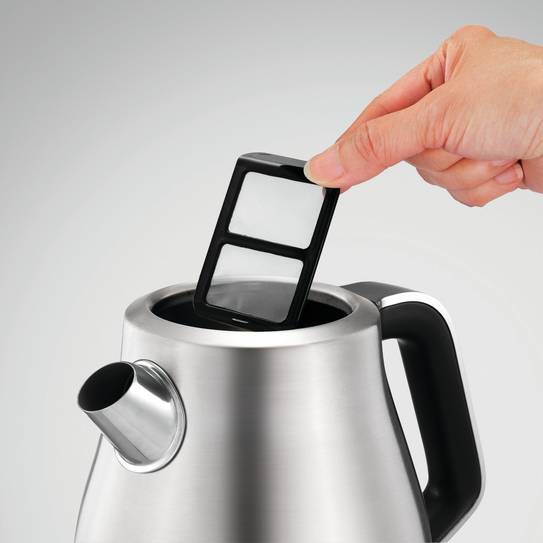 Ręka wyjmuje filtr ze stalowego czajnika. Filtr jest czarny i prostokątny.