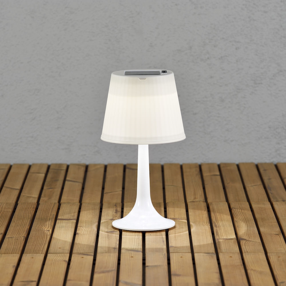 Eine weiße Tischlampe mit Lampenschirm auf einem Holztisch. Die Lampe ist beleuchtet.