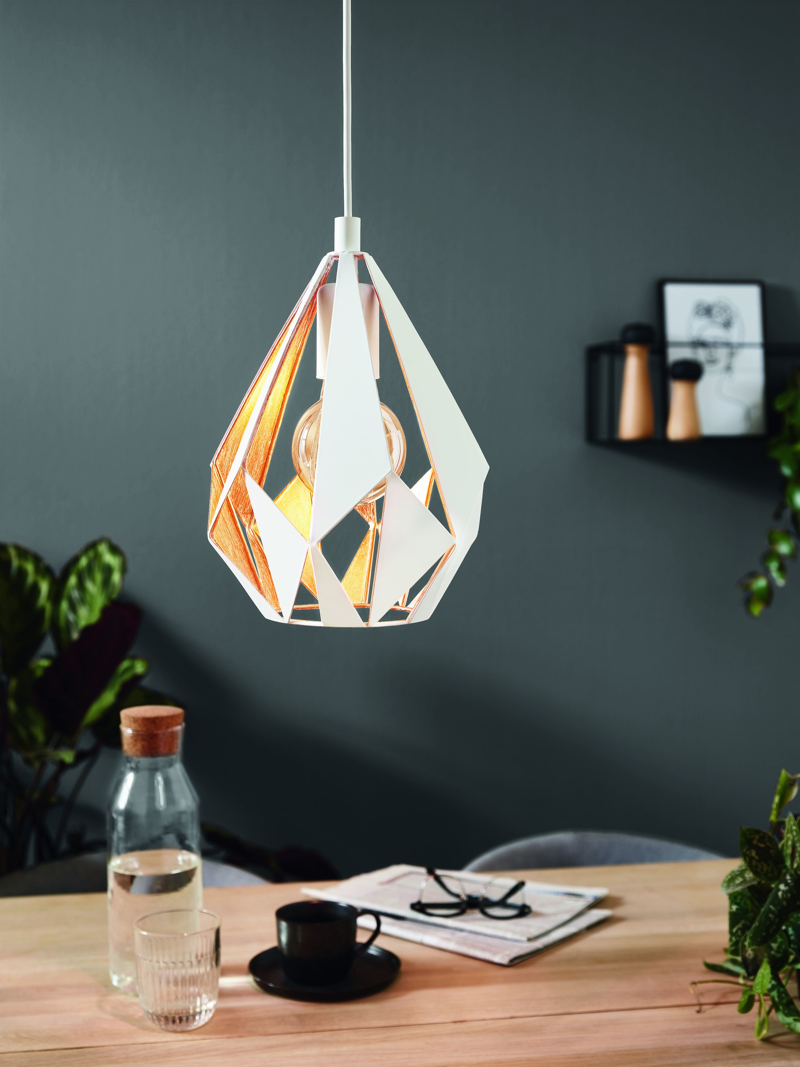 Lampada a sospensione con paralume geometrico sopra un tavolo. È in una stanza. Lo sfondo è grigio.