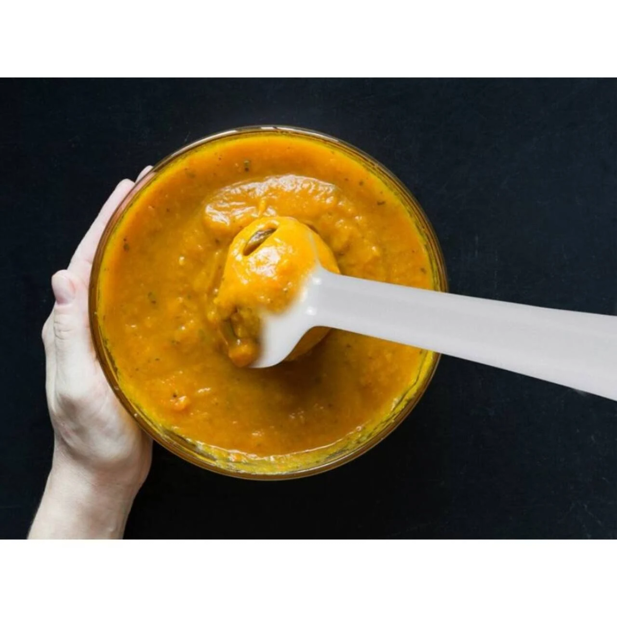 Ręka trzyma biały blender w pomarańczowej zupie, czarne tło.