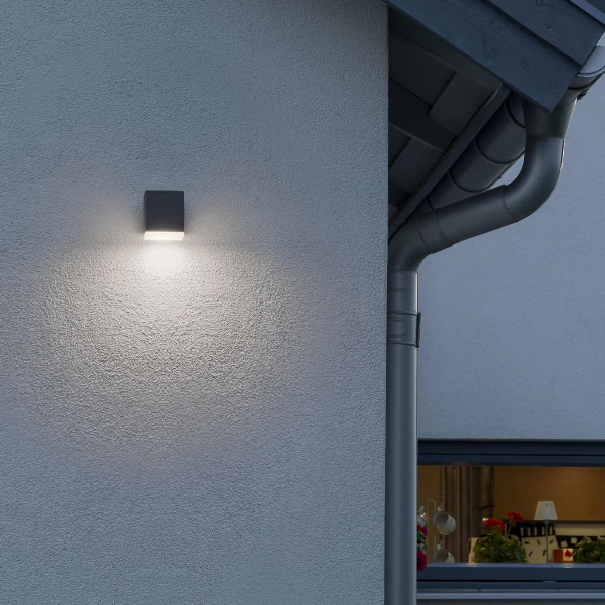 Außenwandleuchte an einer grauen Wand. Die Wandleuchte ist quadratisch und das Licht ist an.