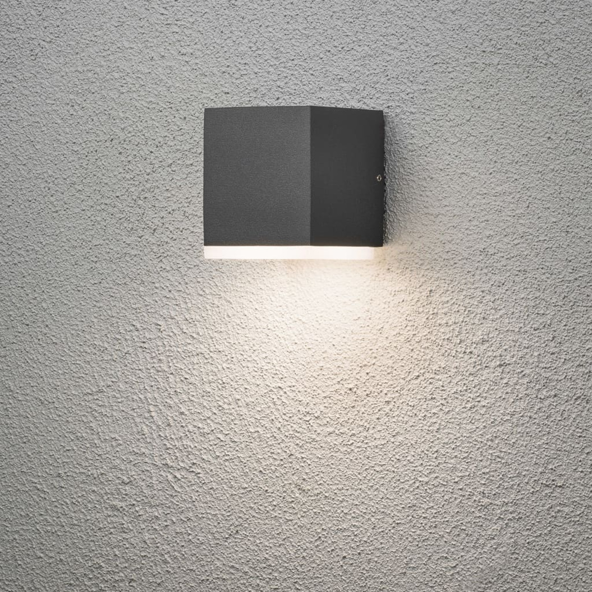 Quadratische Wandleuchte an einer strukturierten Wand. Die Lampe ist dunkelgrau und gibt weißes Licht ab.