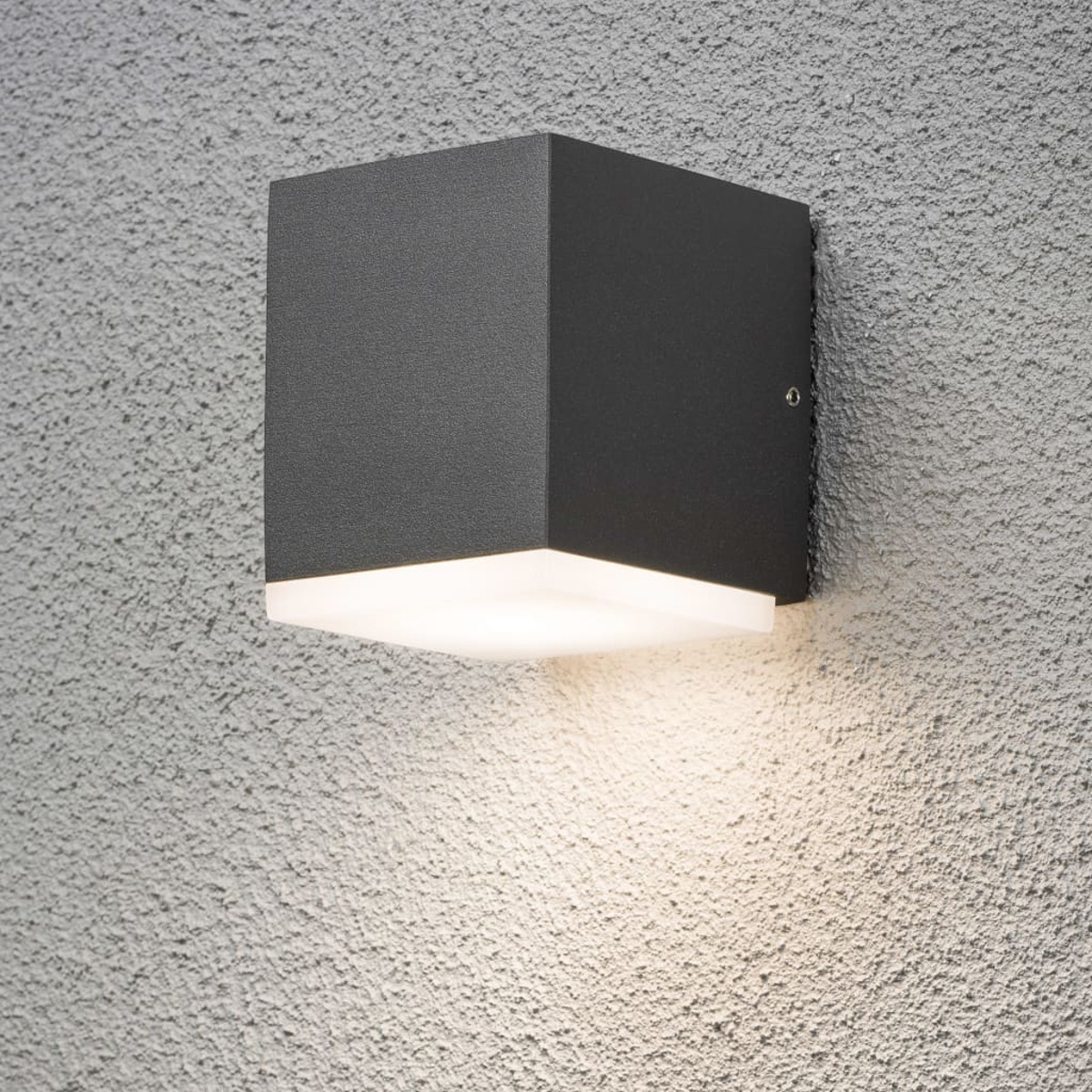 Quadratische Wandleuchte an einer strukturierten Wand. Die Lampe ist dunkelgrau und gibt weißes Licht ab.