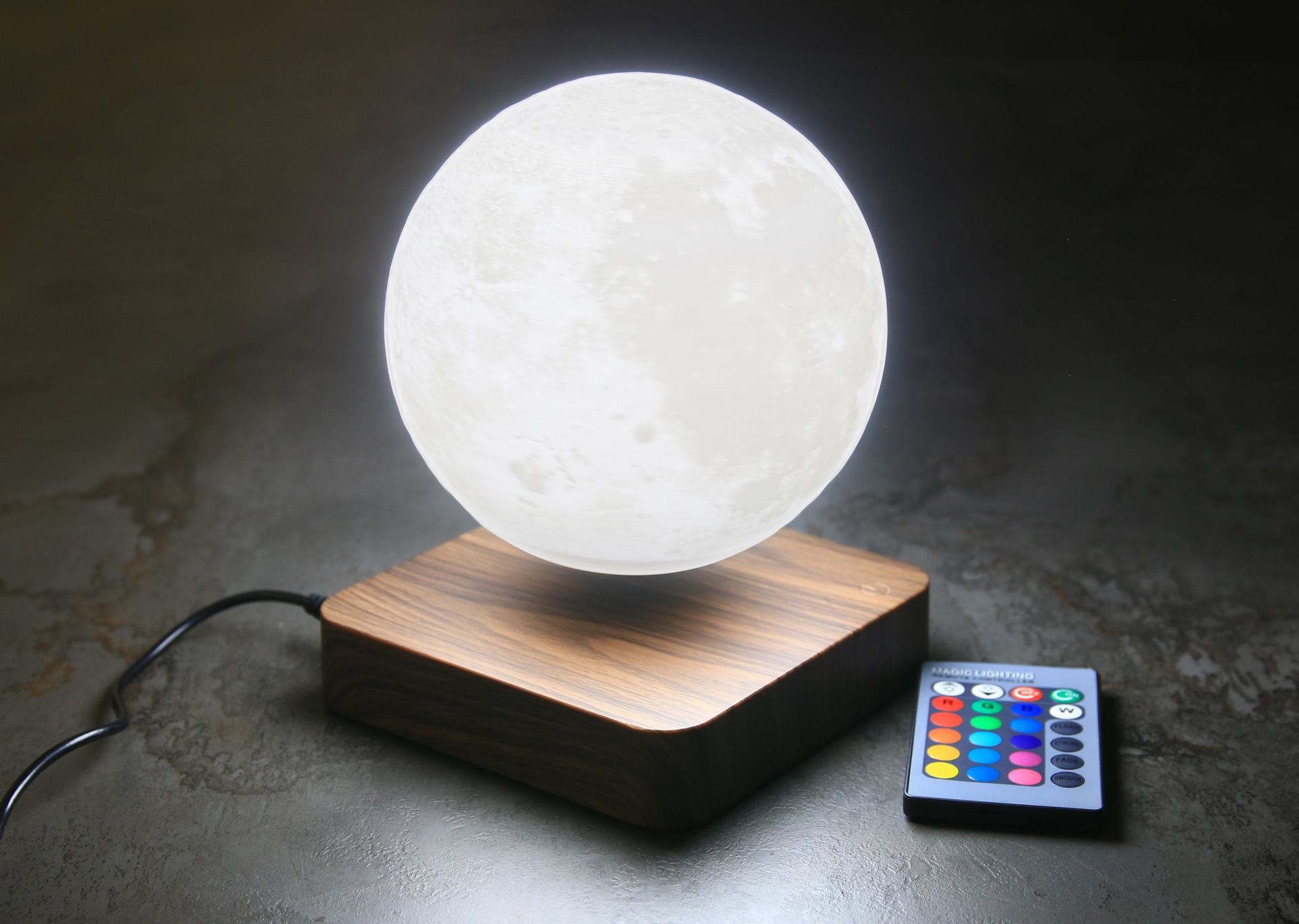 Una lámpara lunar flotante con base de madera y control remoto, sobre una superficie oscura.