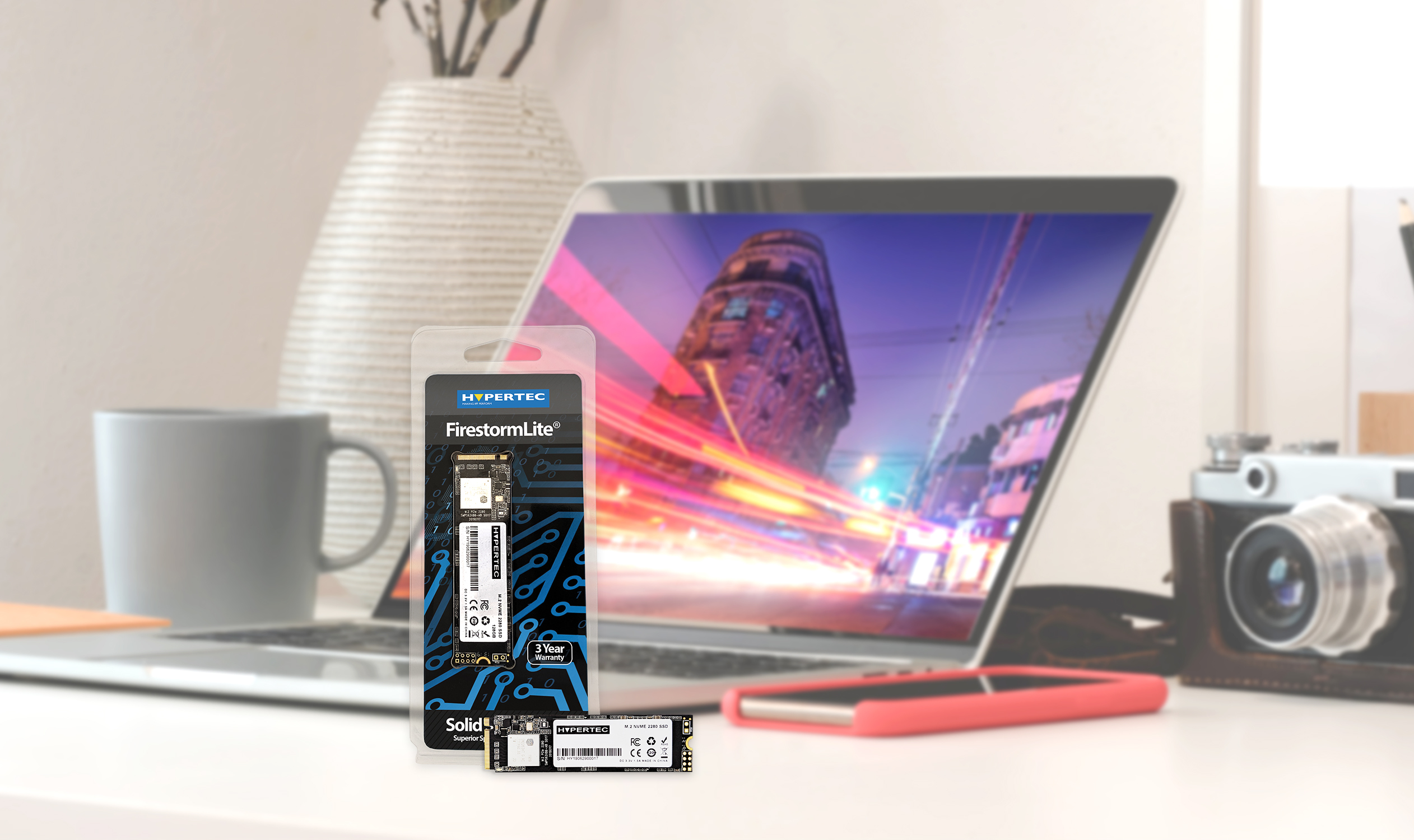 Laptop con confezione SSD davanti. Sfondo urbano sfocato. Altri oggetti sulla scrivania.