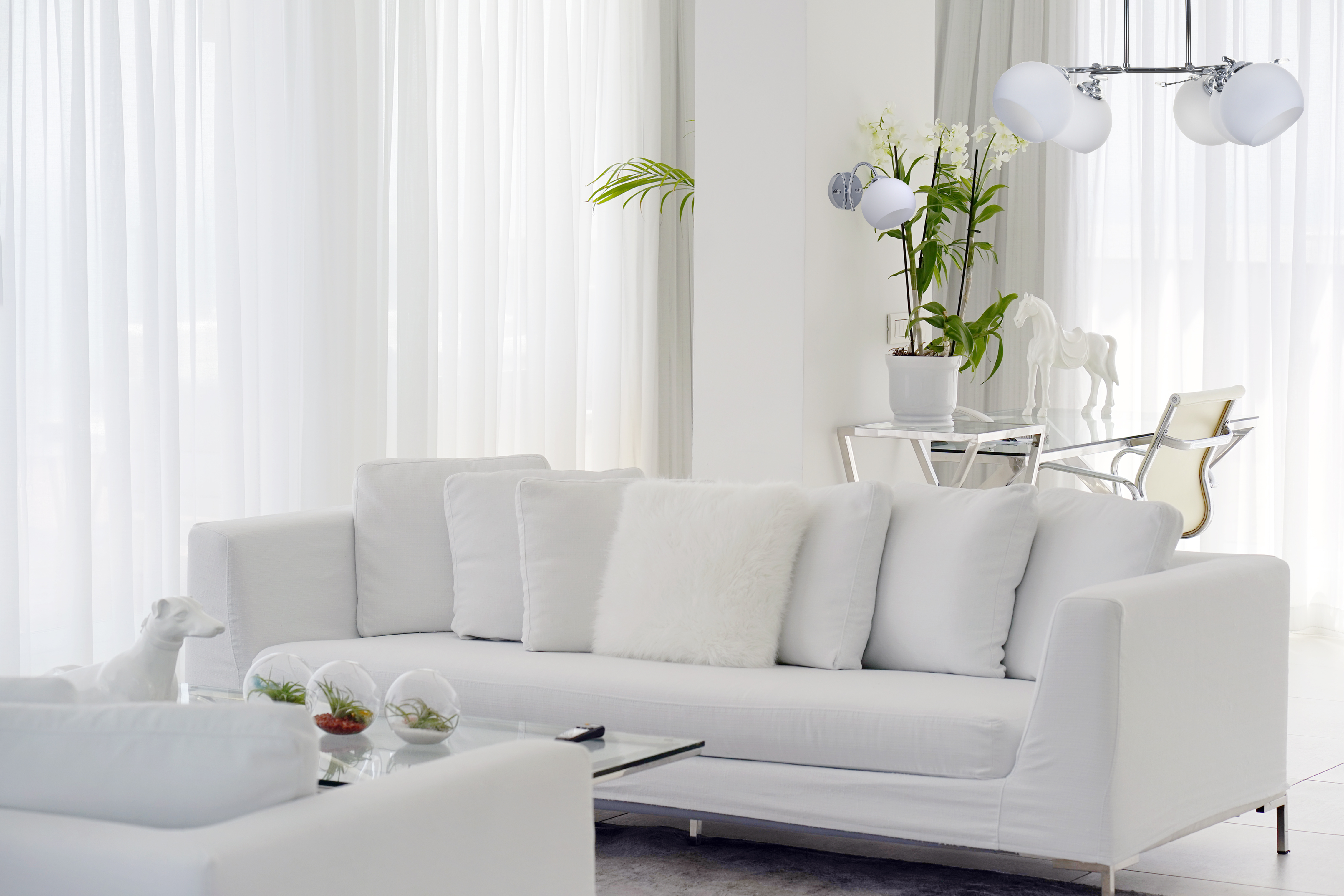Stanza bianca con divano bianco, cuscini e tende trasparenti. Sono presenti oggetti decorativi.