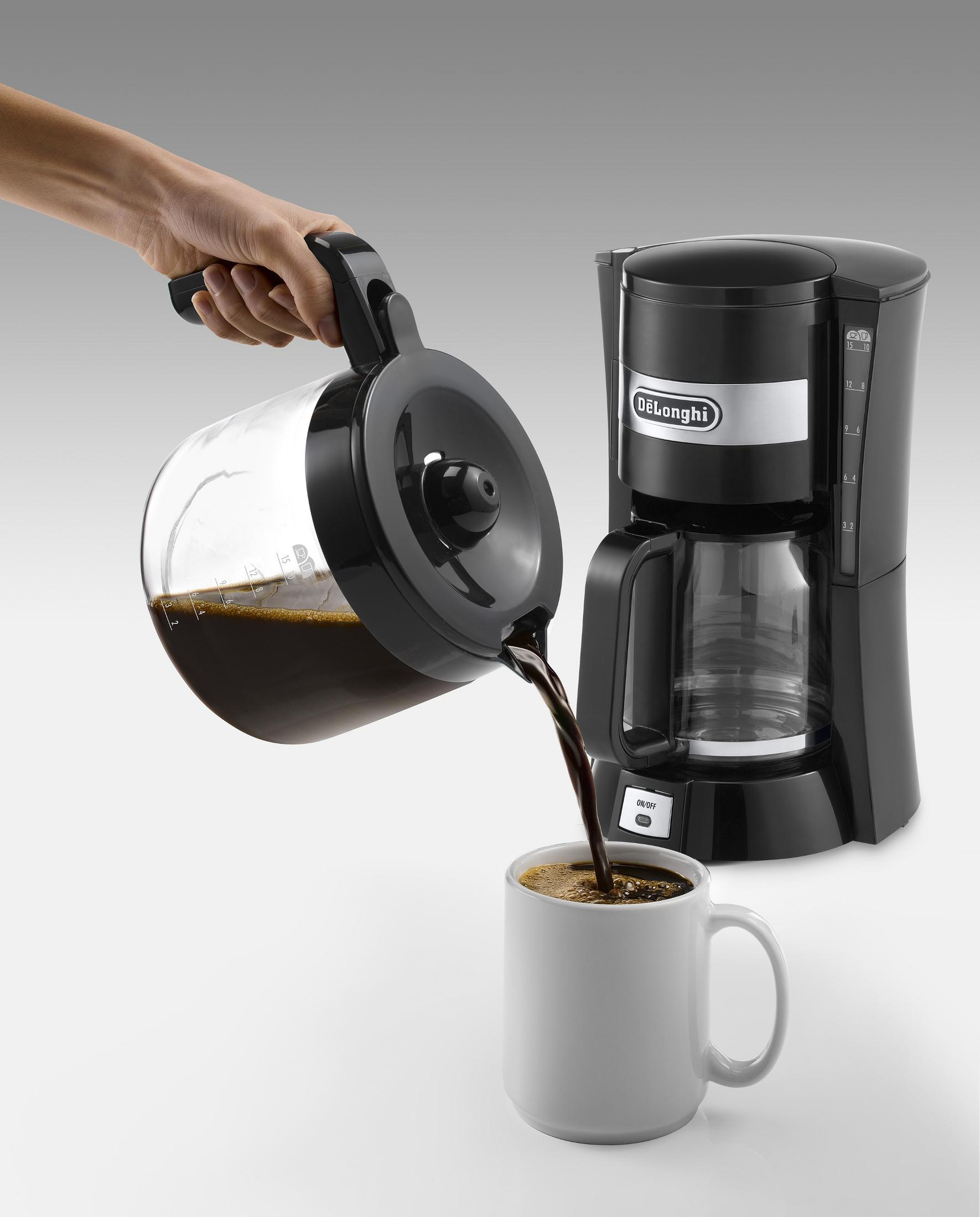 Eine Hand gießt Kaffee aus einer Glaskaraffe in eine weiße Tasse, mit einer Kaffeemaschine.