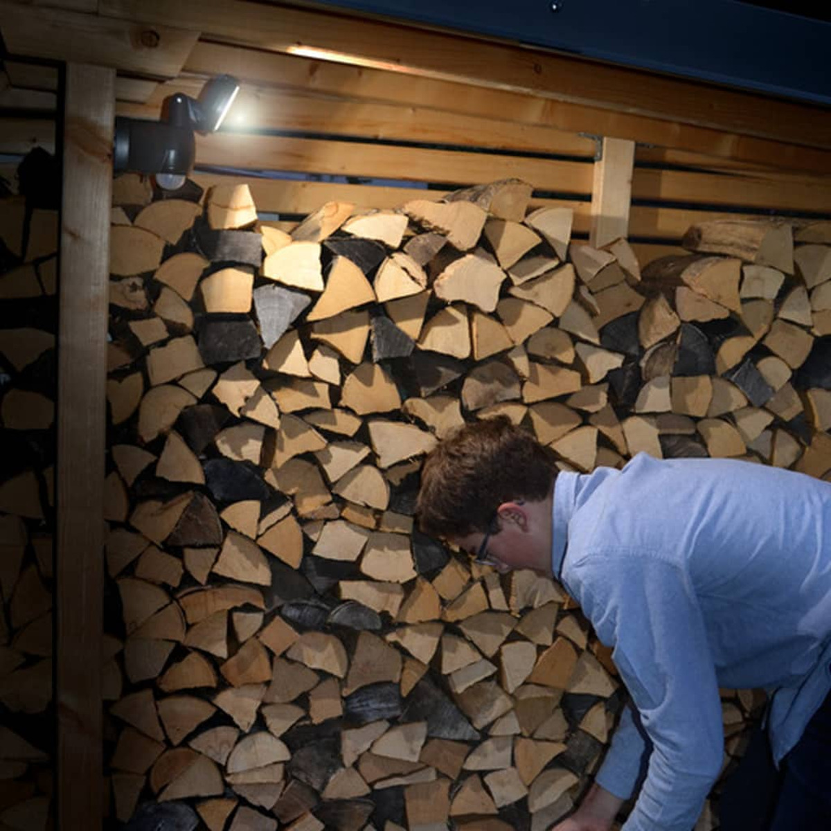 Mężczyzna w pobliżu stosu drewna, oświetlony przez zewnętrzną lampę bezpieczeństwa zamontowaną na drewnianej konstrukcji.