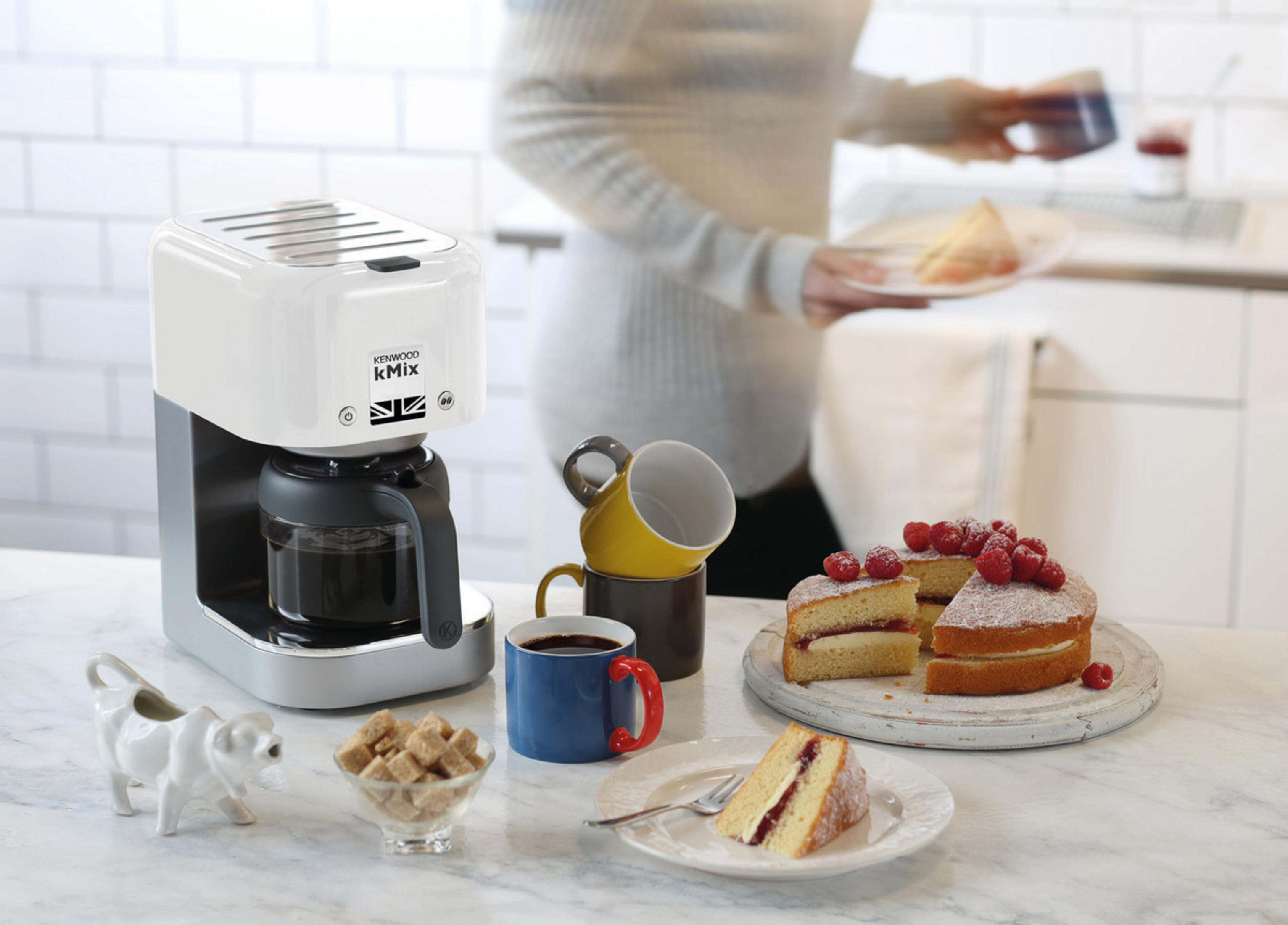 Scena kuchenna z białym Kenwood KMix. Kawa, ciasto, owoce, kubki. Osoba w tle trzymająca tacę.