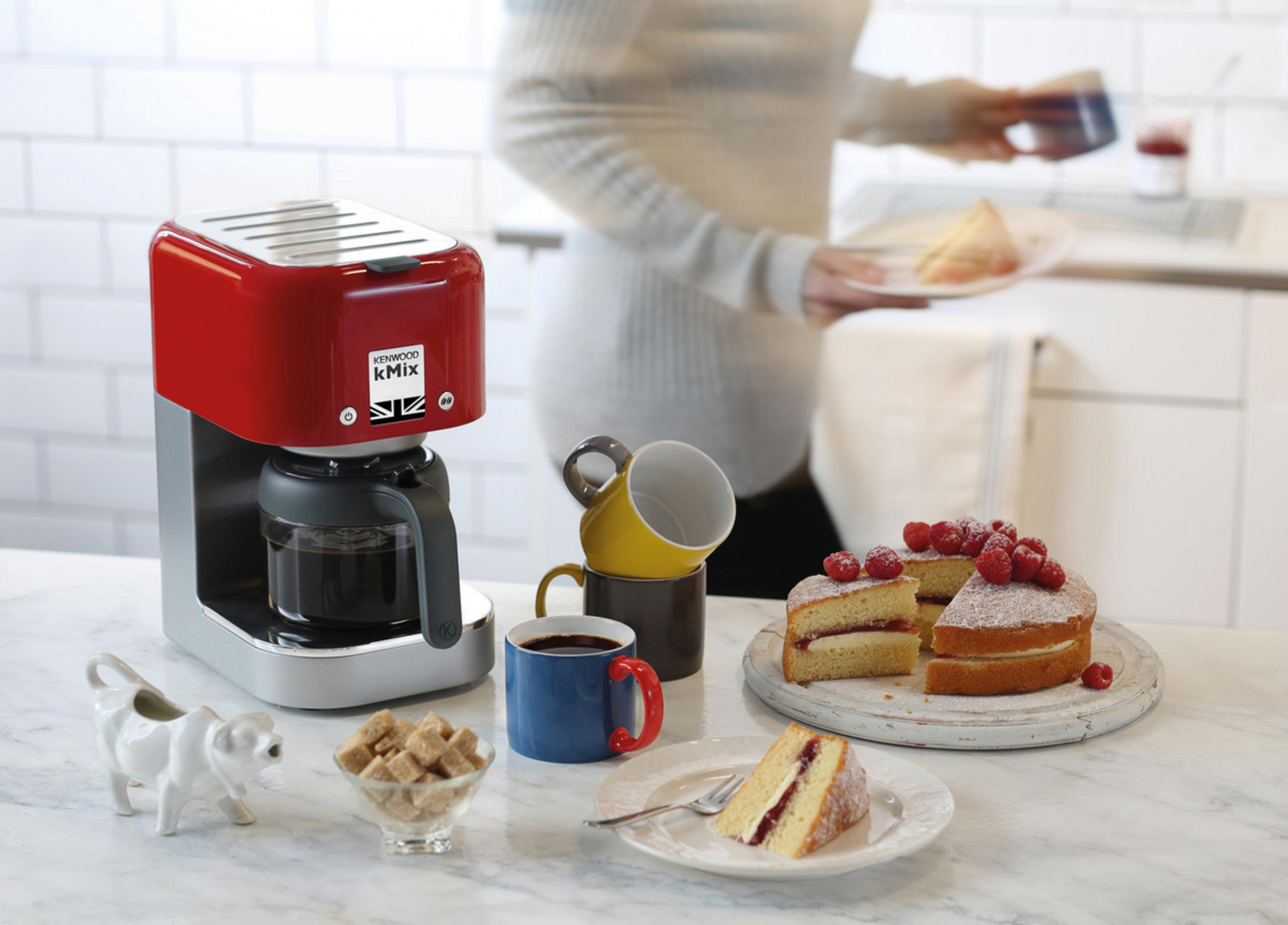 Czerwony ekspres do kawy z kubkami, ciastem, cukrem i kobietą w tle.