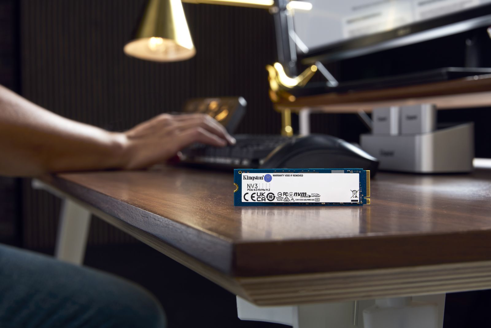 SSD en escritorio, usuario tecleando, lámpara de escritorio. Kingston NV2 visible.