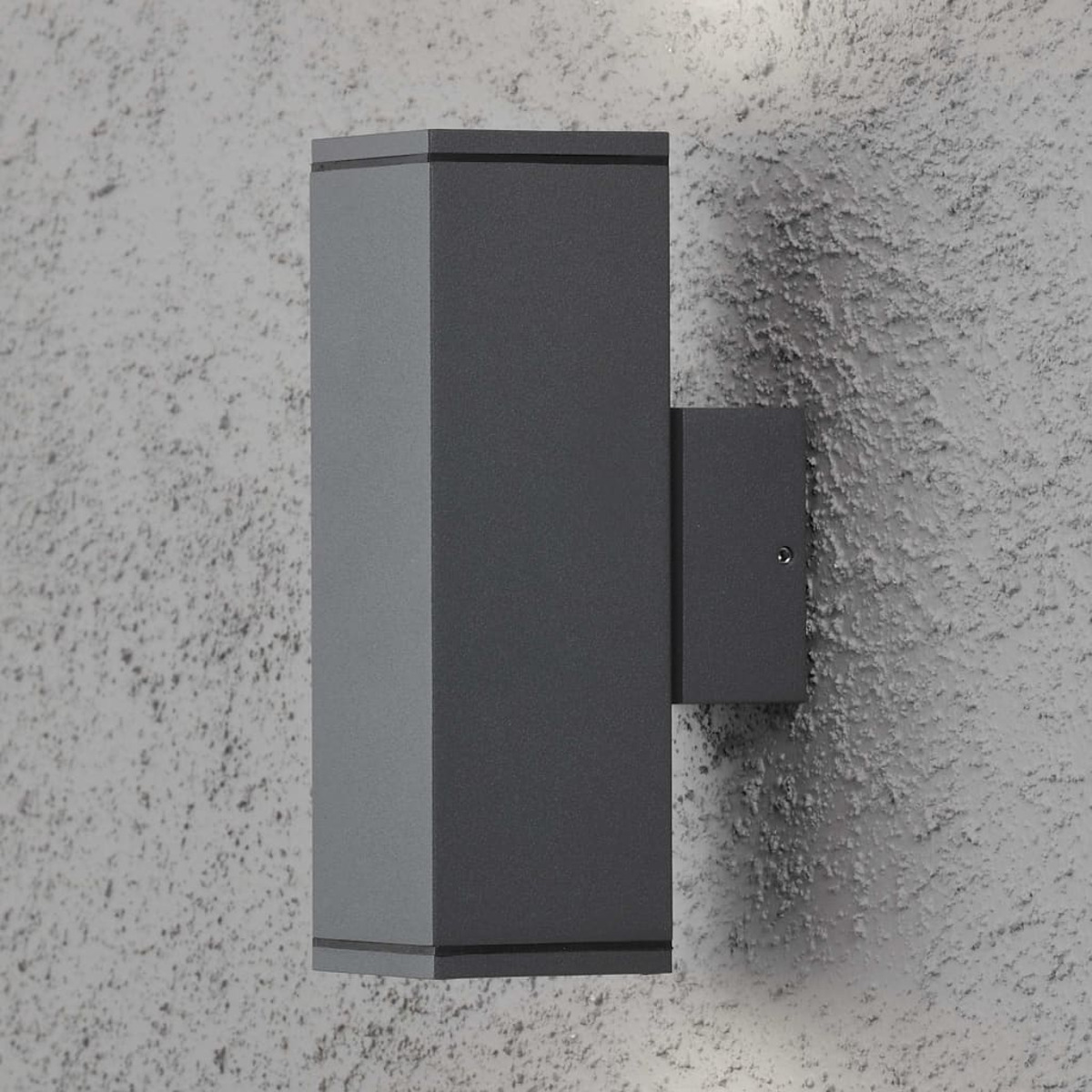Una lampada da parete esterna grigio scuro su un muro strutturato, con una luce simile sfocata sullo sfondo.