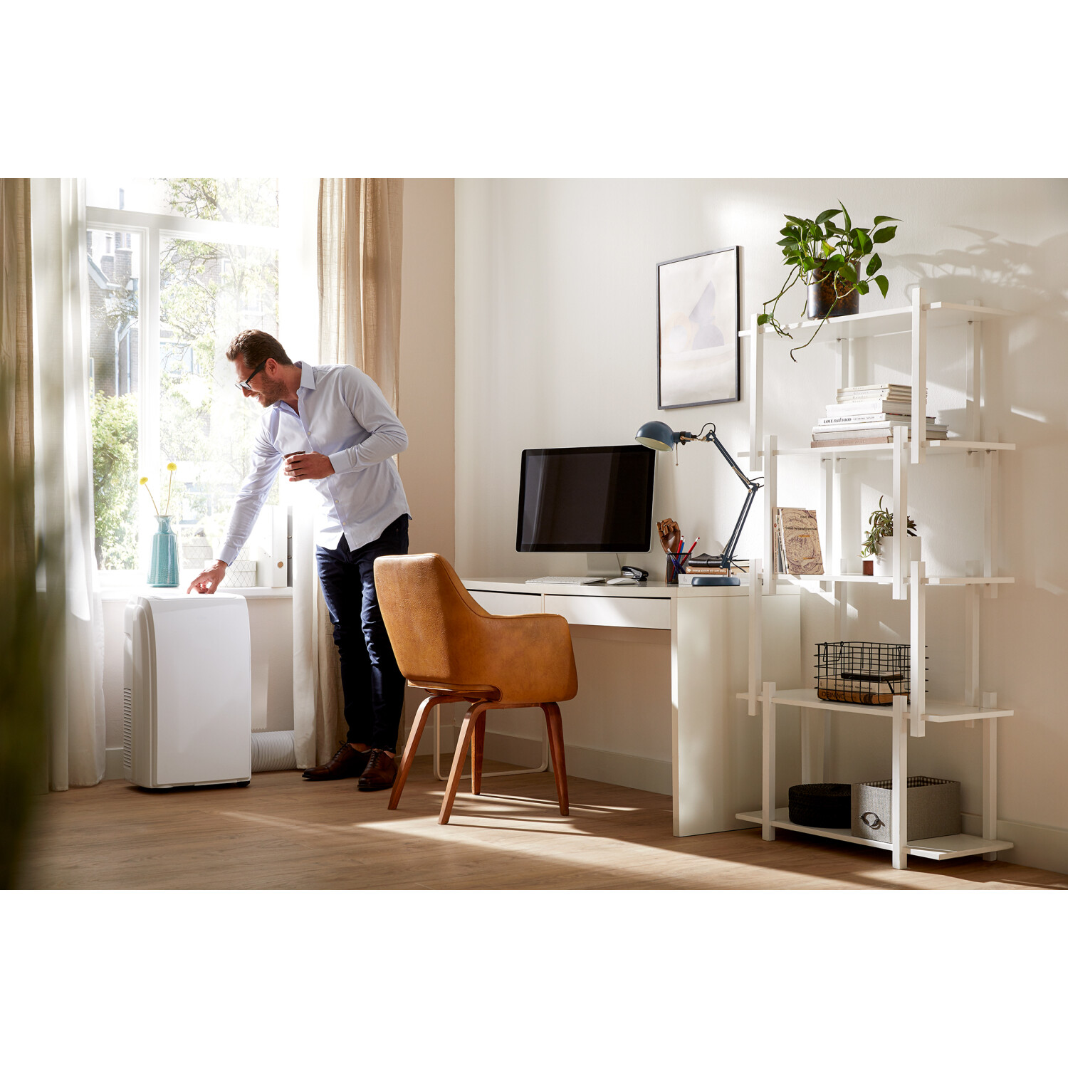 A man in a room, by a window, next to a white air conditioner and a desk with a computer.