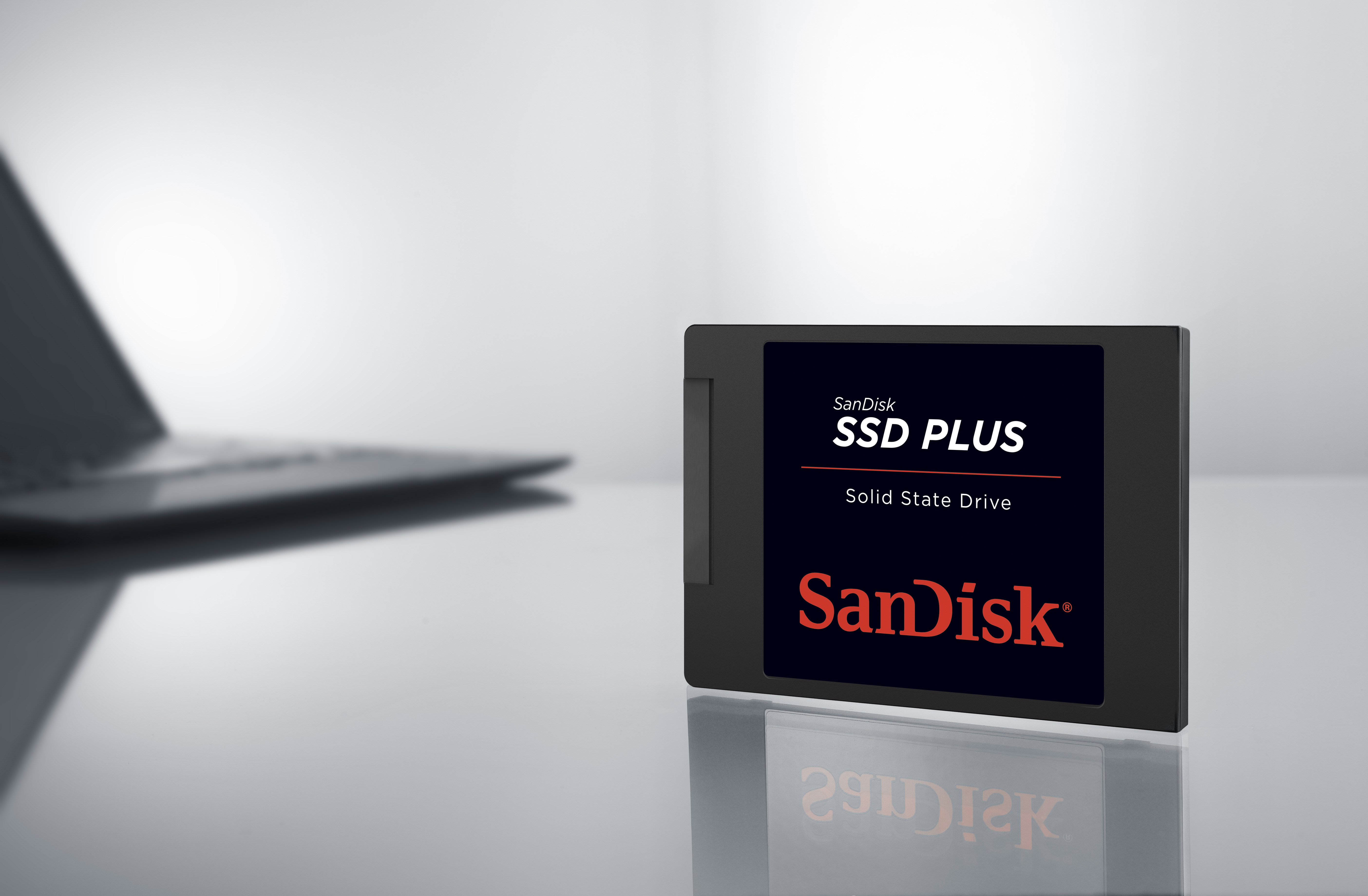 Eine schwarze Sandisk SSD Plus auf weißer Oberfläche, neben einem Laptop. Der Hintergrund ist weiß.