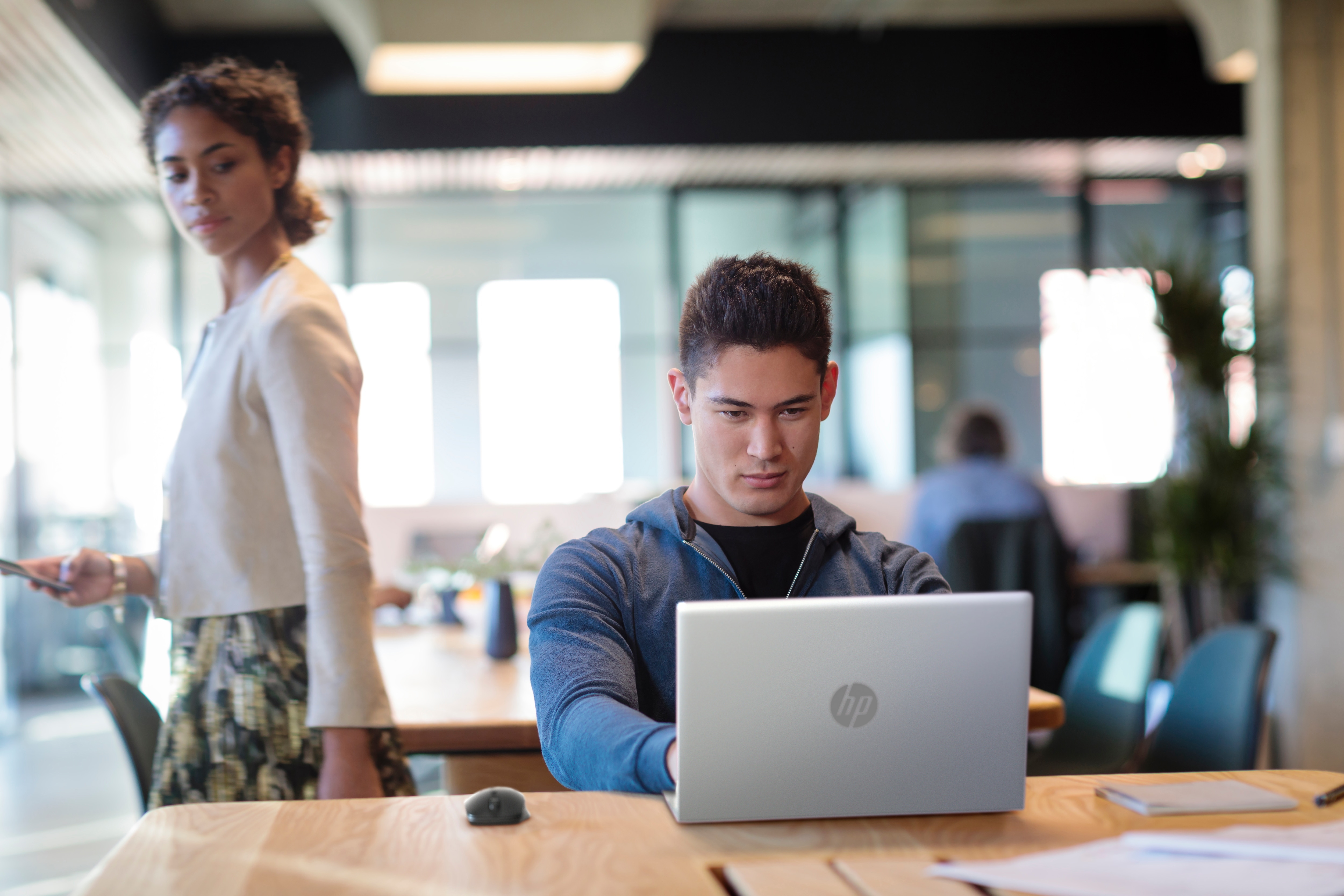 A man using a laptop at a table. A woman stands in the background. Office setting.