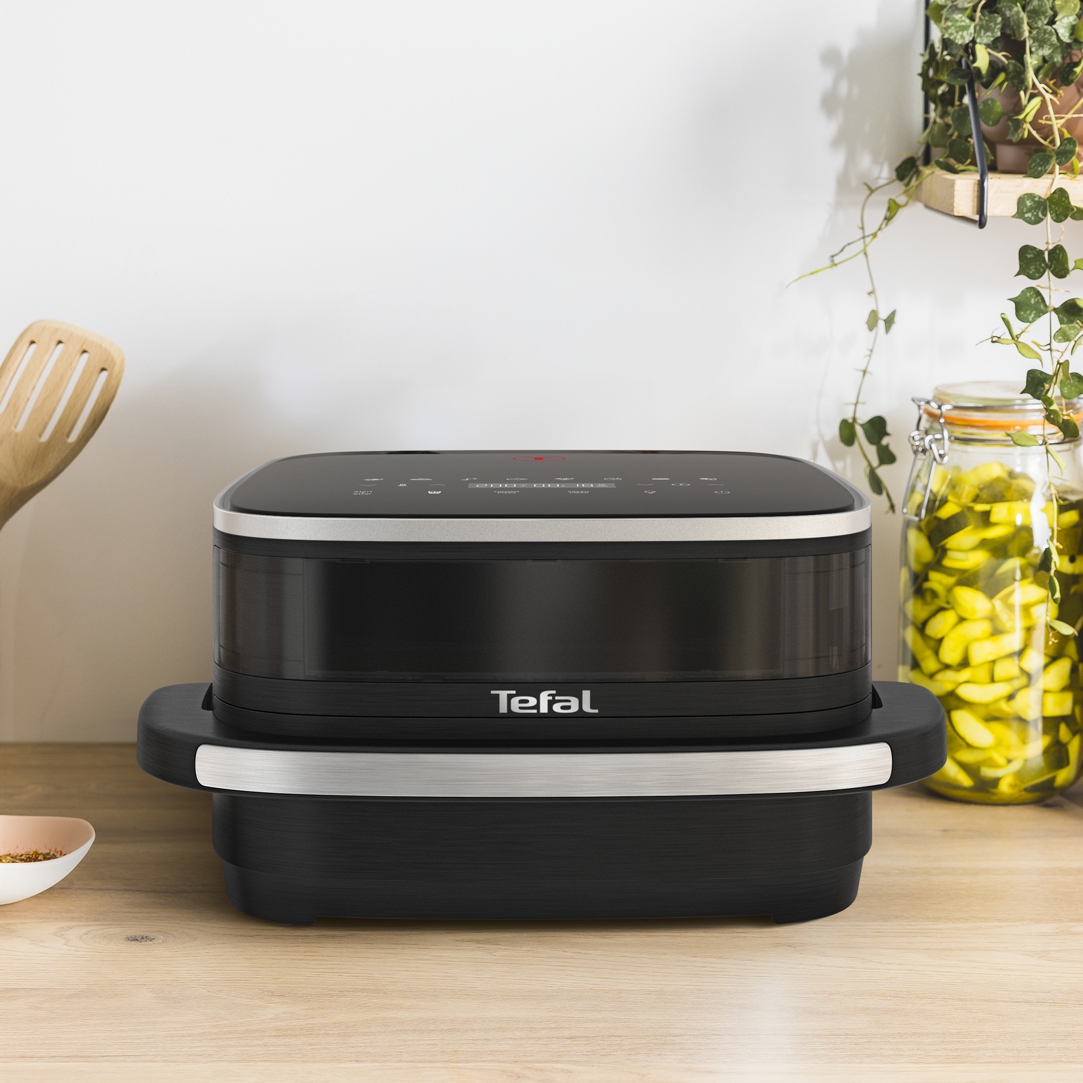 Czarna frytownica powietrzna na drewnianej powierzchni. Widoczne logo Tefal. Tło: kuchnia z roślinami i słoikami.